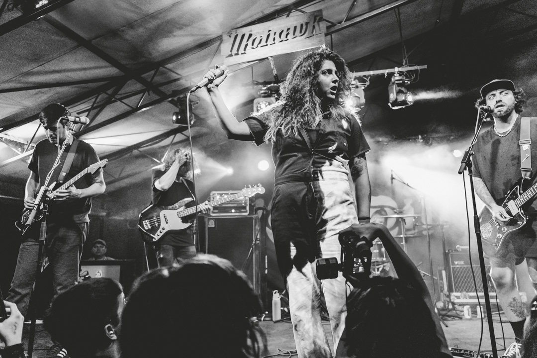 A woman performs on stage with a band, holding a microphone, in a black and white photo at a live music event.
