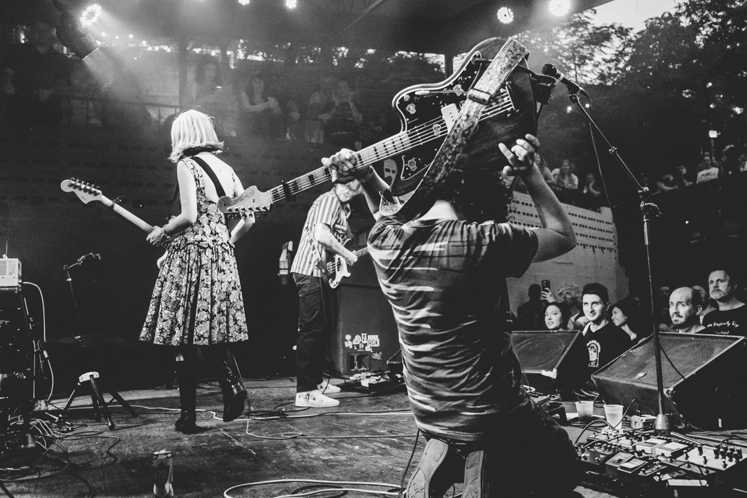 Musicians perform on stage with guitars and a keyboard, audience watching in the background, black and white photo.
