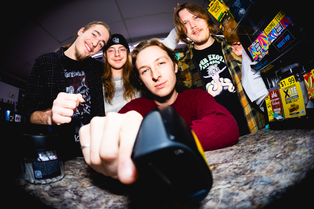 Four young people at a counter, one taking a selfie with a camera, smiling and posing. Store shelves with snacks visible.