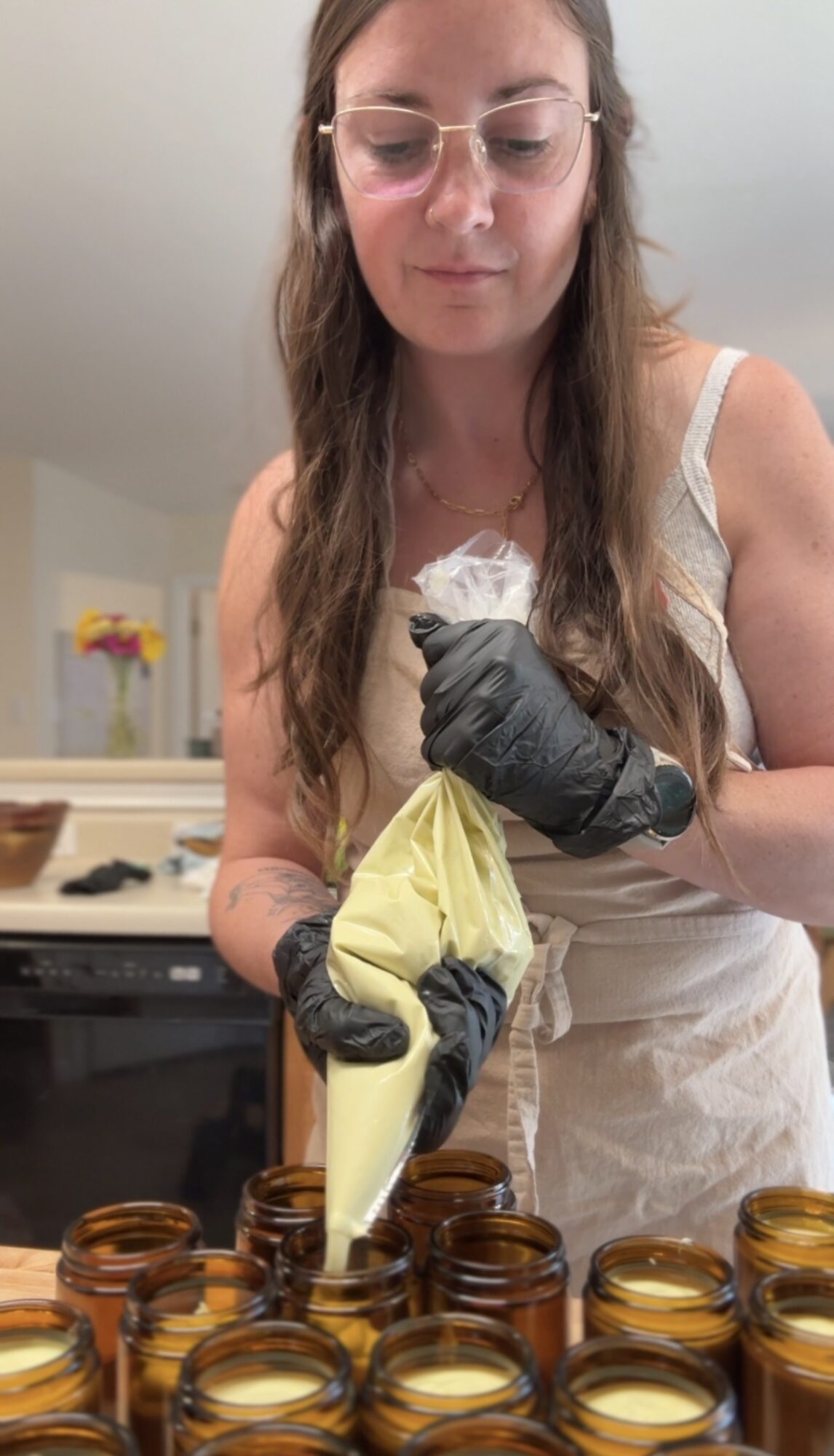 Woman wearing glasses and gloves, pouring wax into small jars, with a flower arrangement in the background.