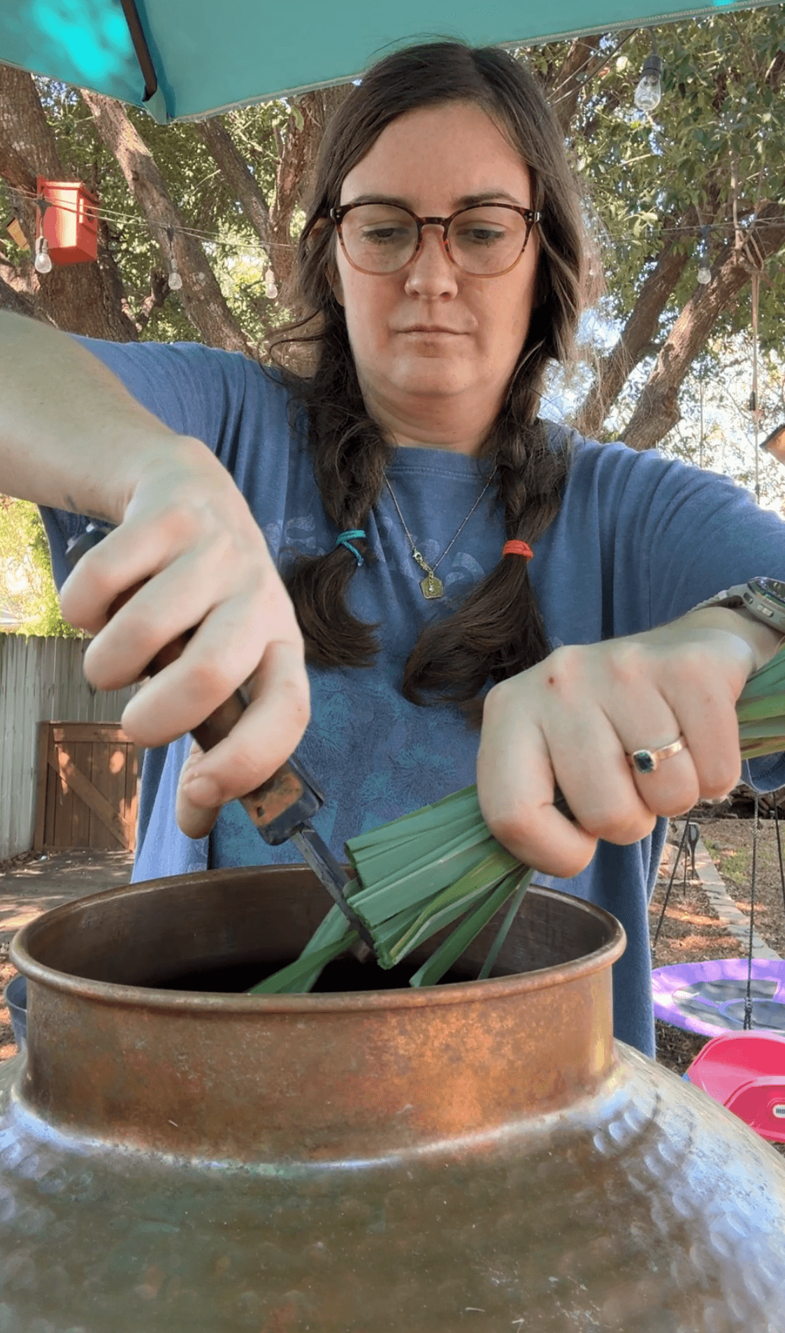 Woman wearing glasses and gloves, stirring a large pot outdoors with trees in background.