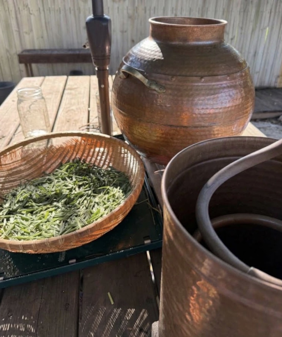 Clay pot, basket with green herbs, glass, and black container on wooden table outdoors.