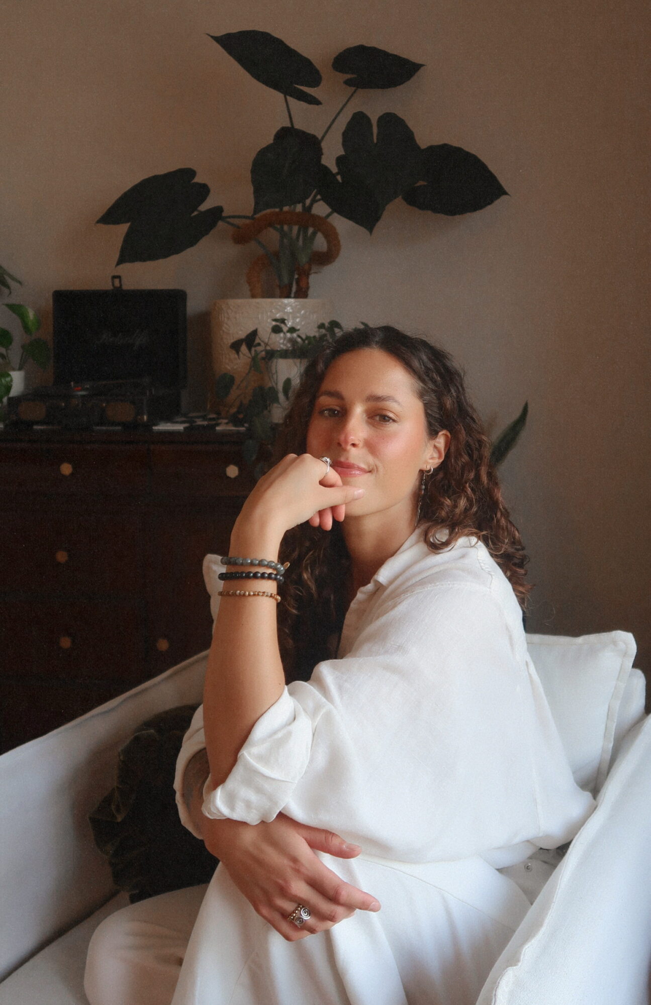 Woman with curly hair sitting on a white couch, wearing a white outfit, with a plant and furniture in the background.