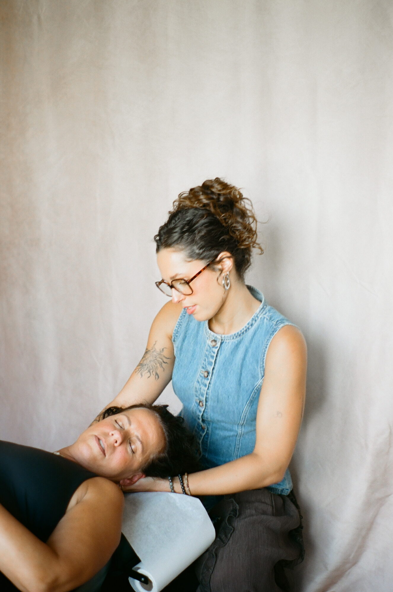 Two women, one with glasses and curly hair, gently touching the other's head, against a plain background.