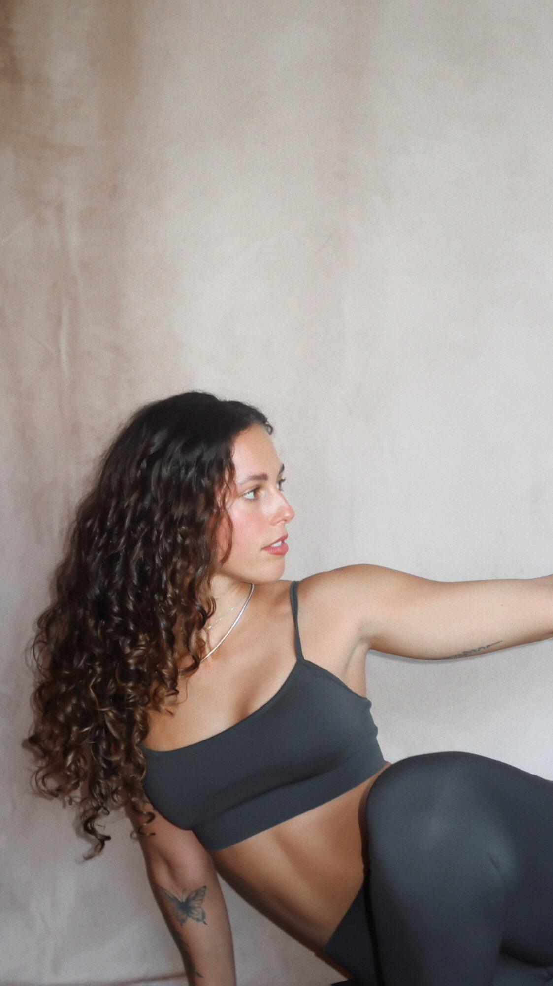 Woman with curly hair in a black sports bra and leggings, sitting against a plain wall, extending her arm.