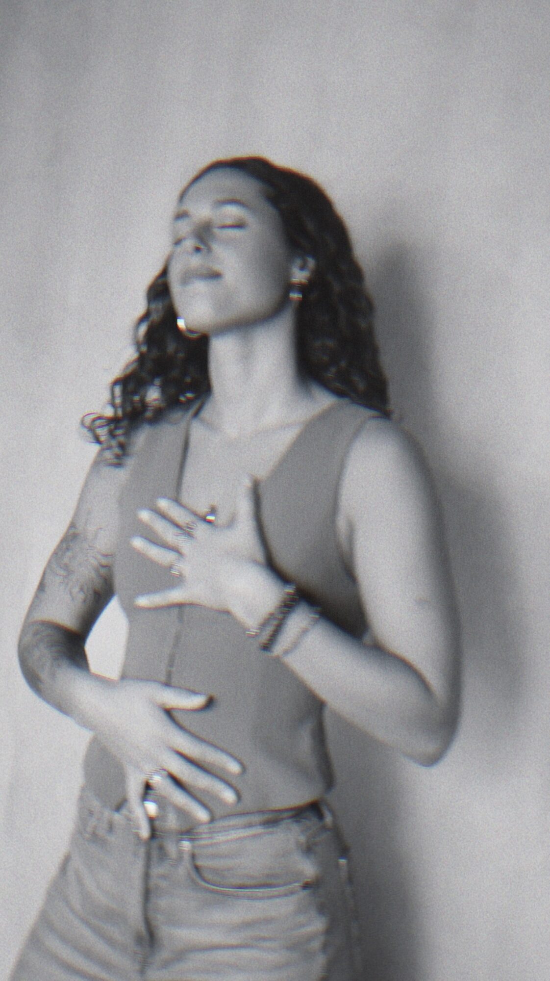 Woman with curly hair, eyes closed, hands on chest and stomach, standing against a plain wall.