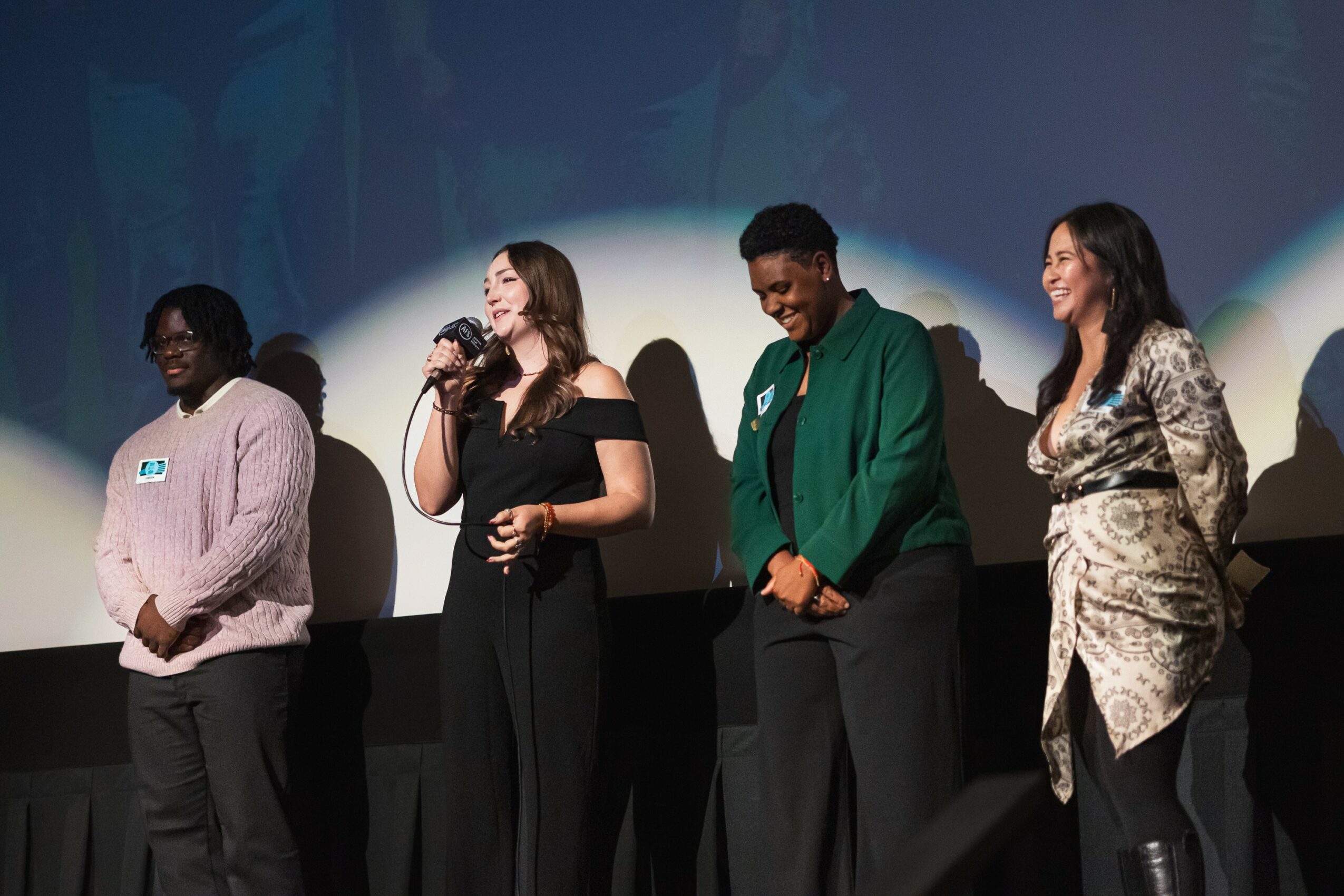 Four people standing on stage, one speaking into a microphone, with a dark background and spotlight. Group appears to be at an event.