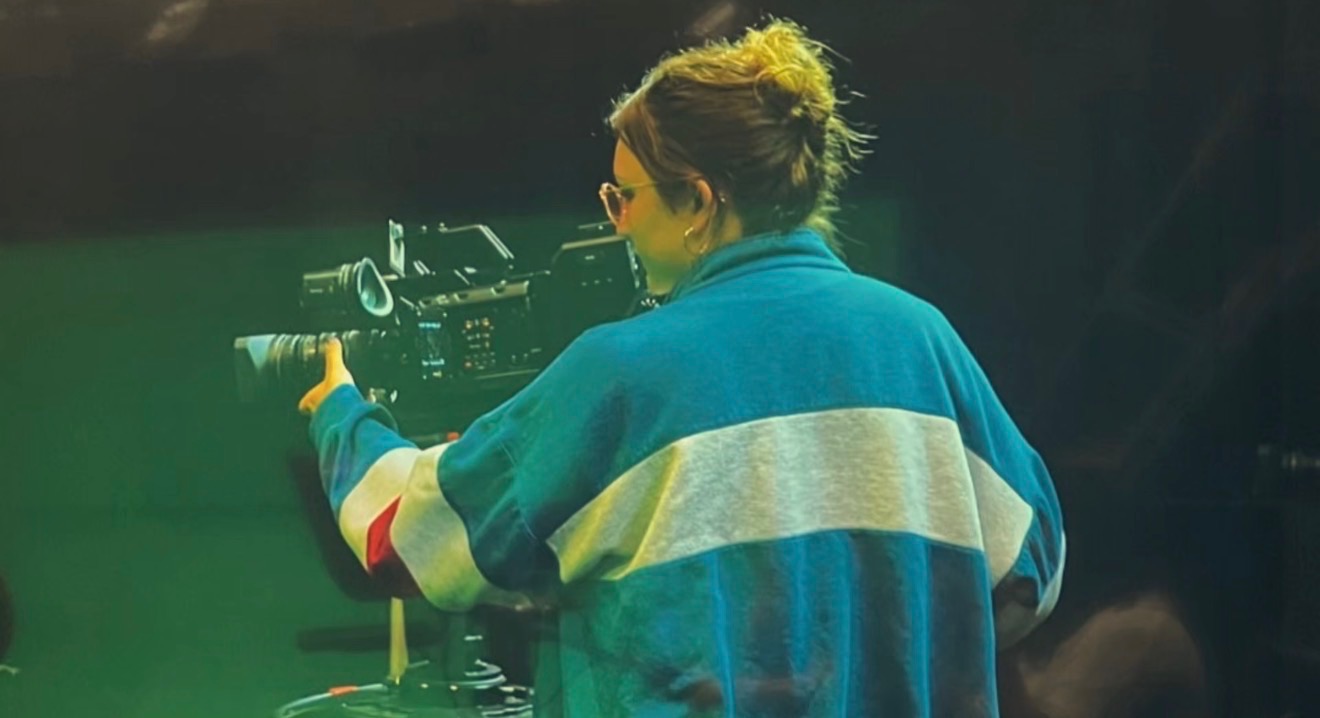 Person with short hair wearing a blue and white jacket, operating a camera or filming equipment, facing away from the camera.