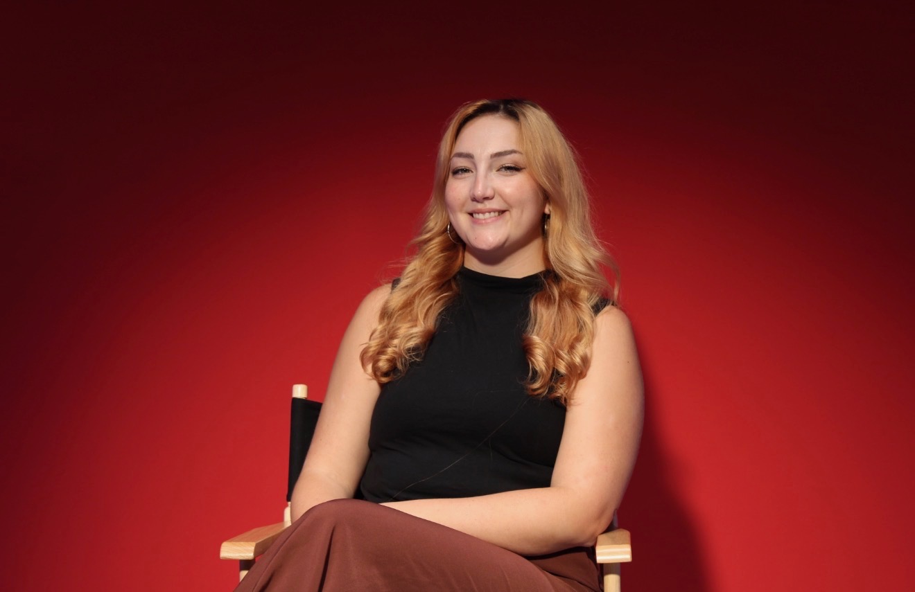 Young woman with long wavy hair smiling, sitting on a chair against a red background.