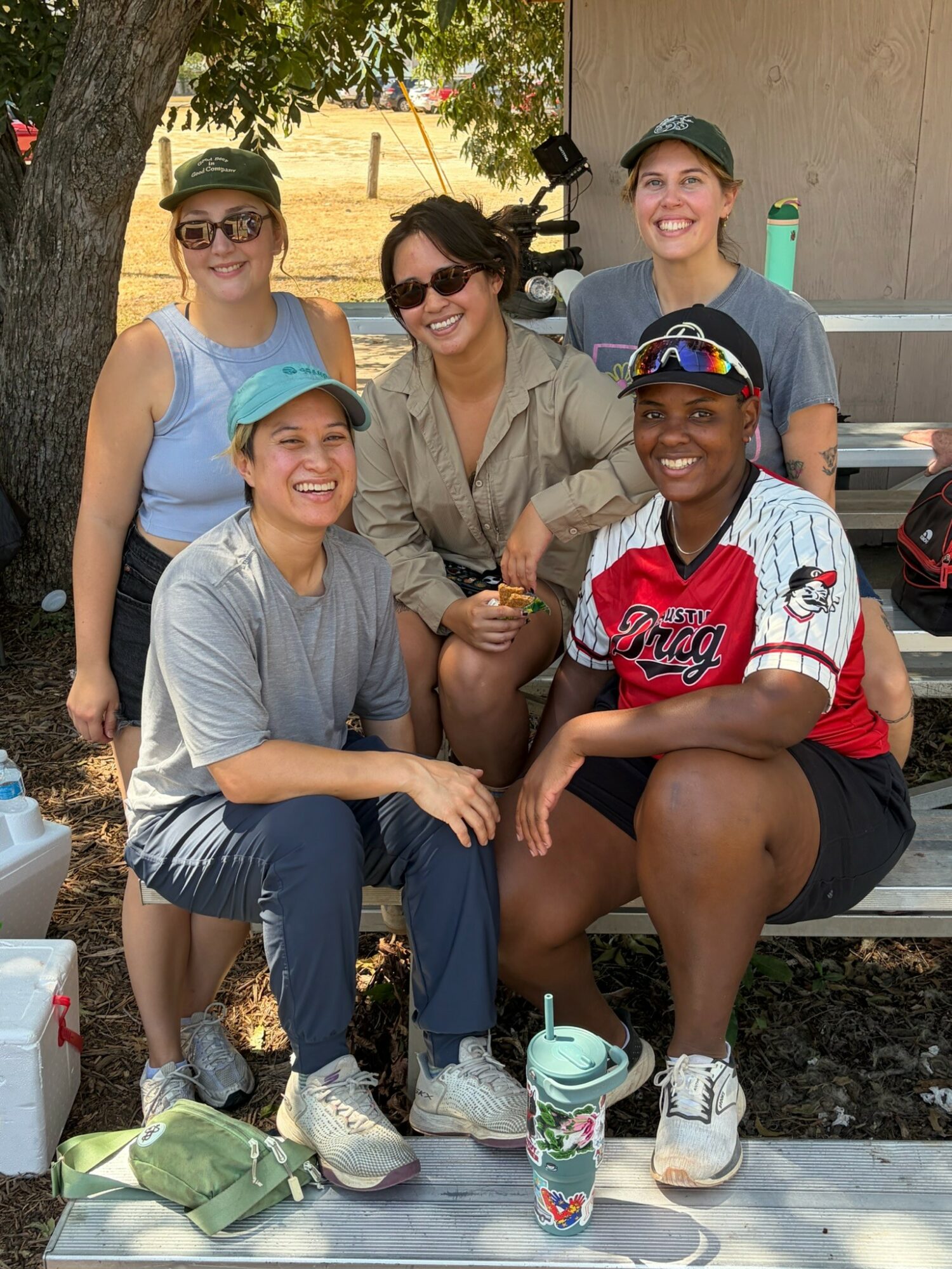 Five women outdoors, smiling, sitting and standing near a tree and a building, with a picnic table and drinks.