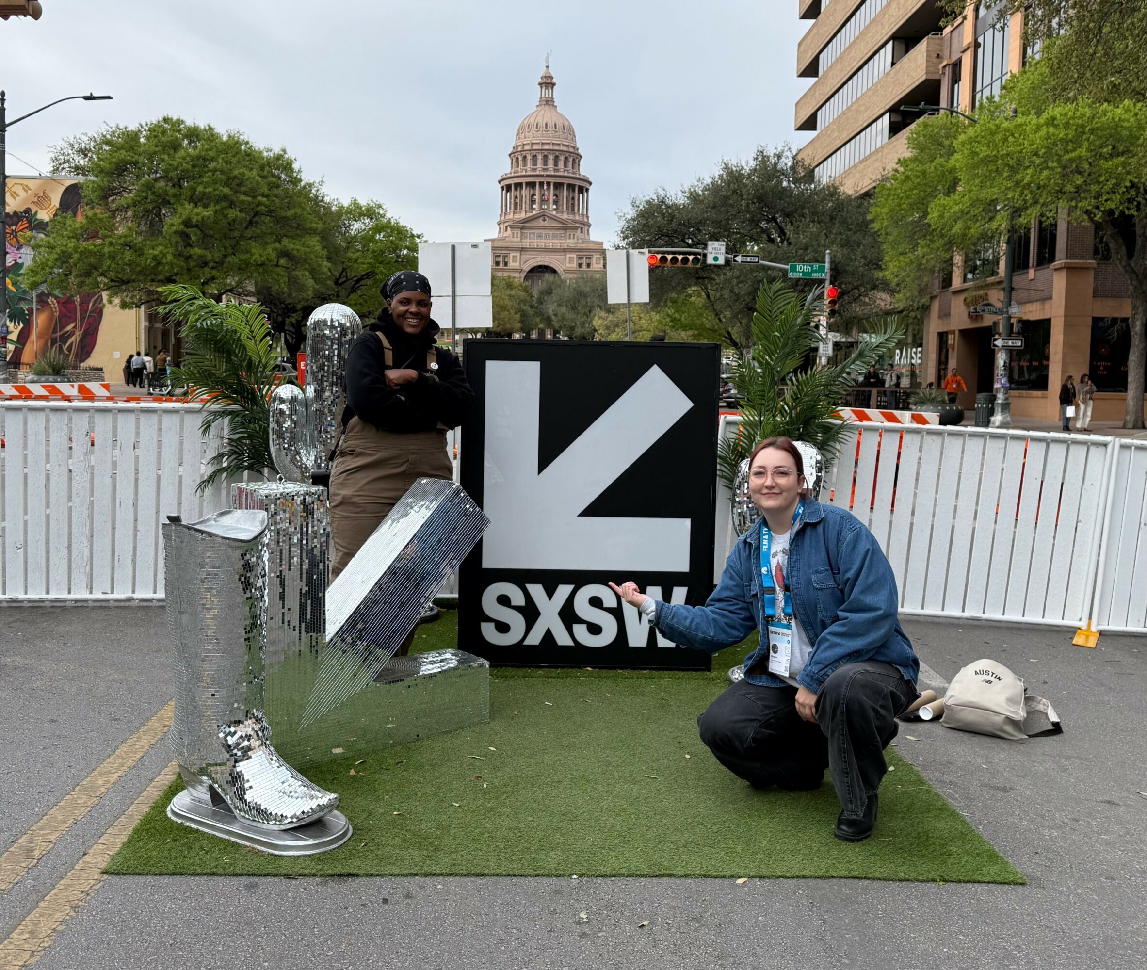Two people pose next to a large SXSW sign on a street with trees and buildings, with the Texas State Capitol in the background.