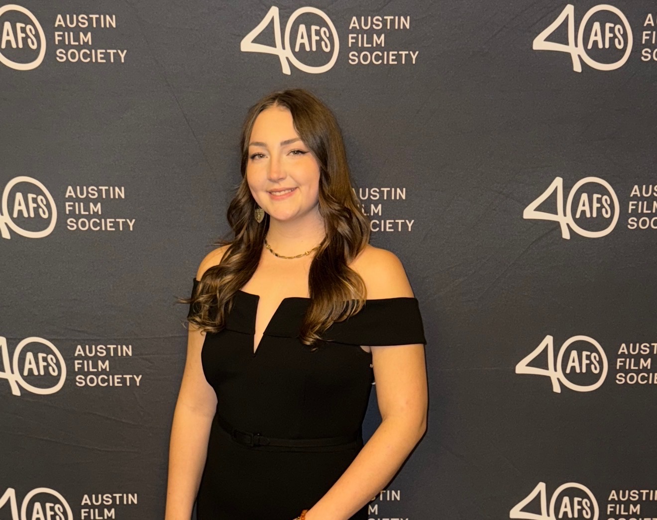 Young woman with long wavy hair in a black off-shoulder dress smiling at an event backdrop.