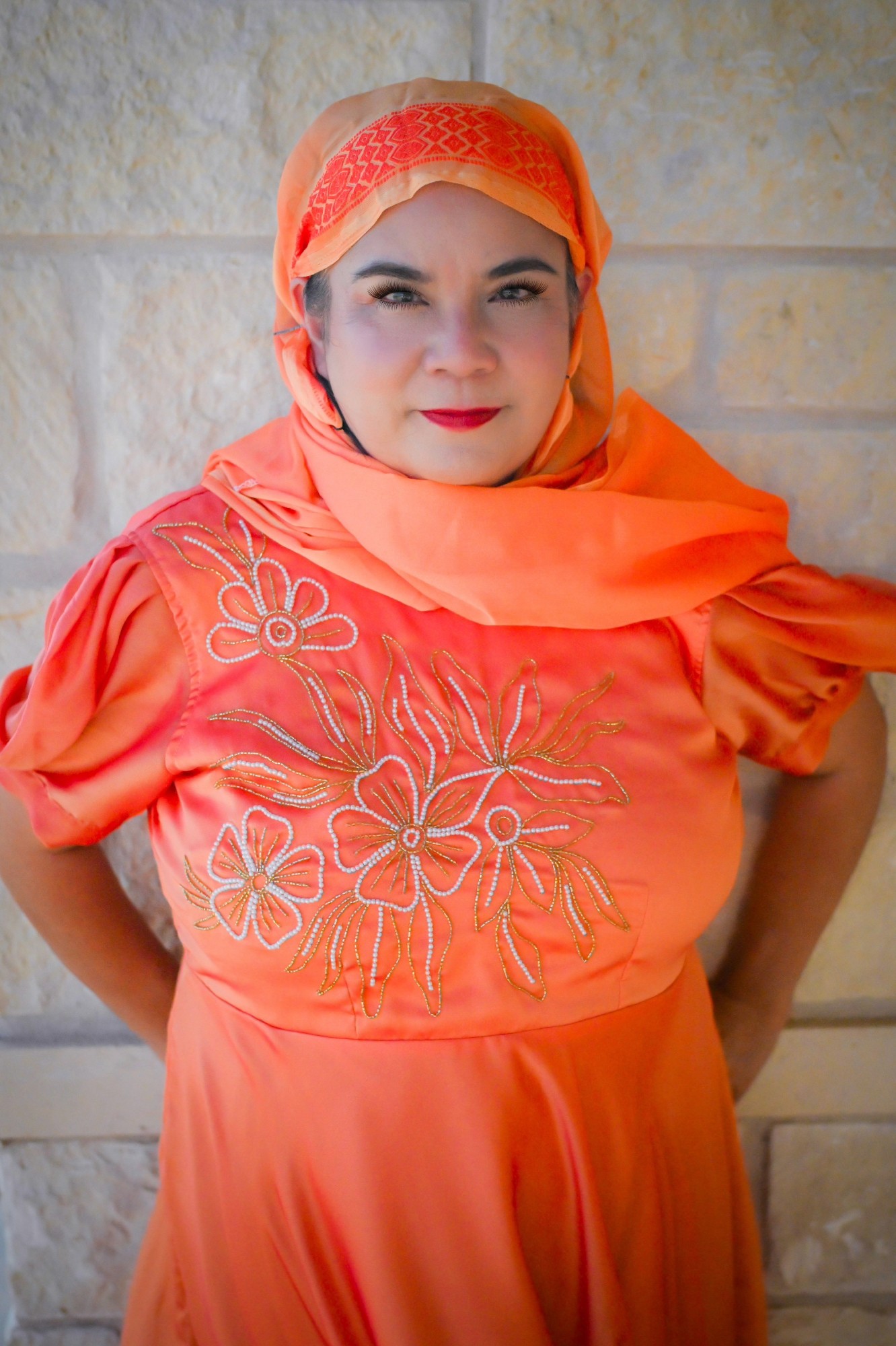 Woman wearing an orange headscarf and dress with floral embroidery, standing against a stone wall.