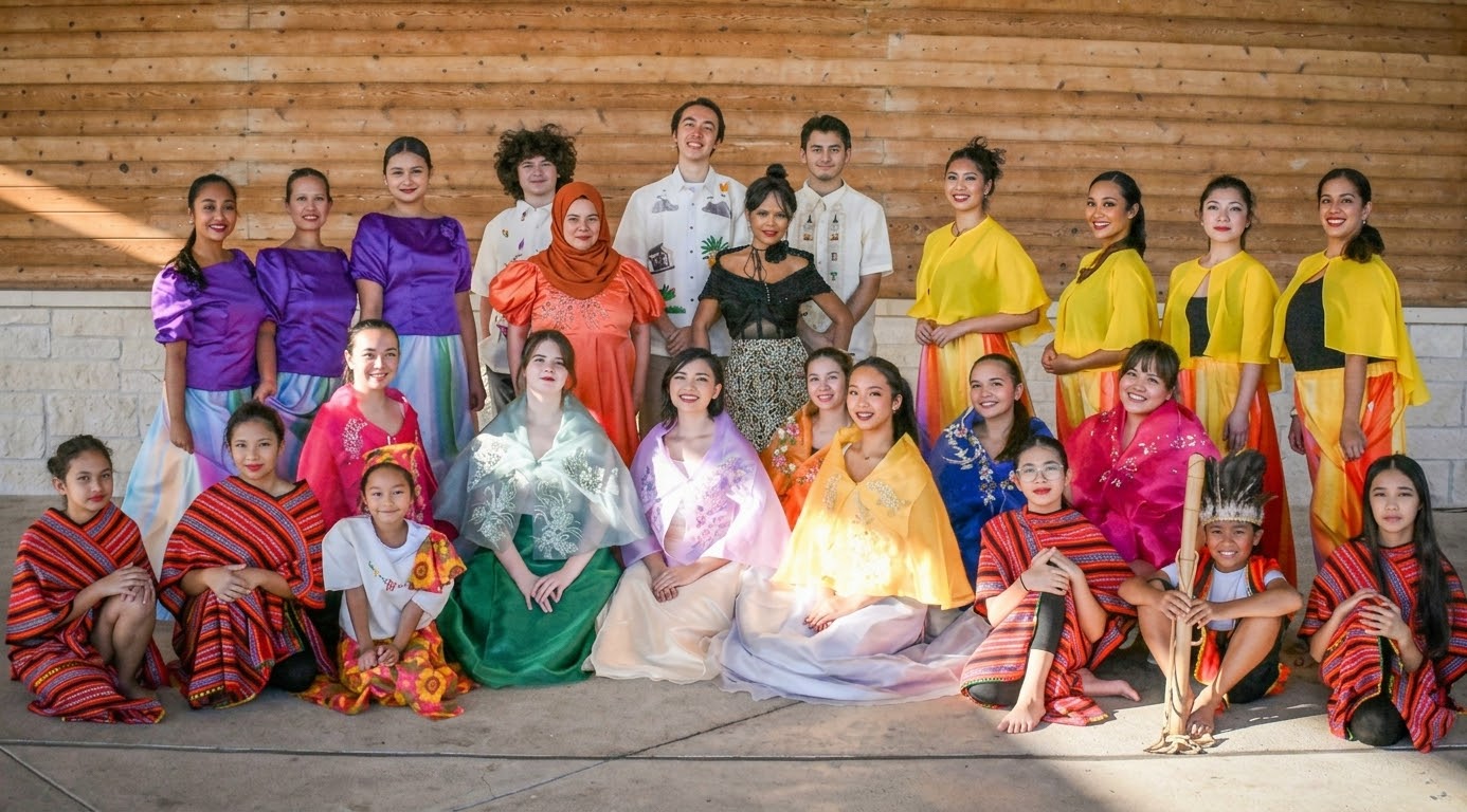 Group of children in colorful costumes on wooden stage, some sitting and some standing, smiling at the camera.