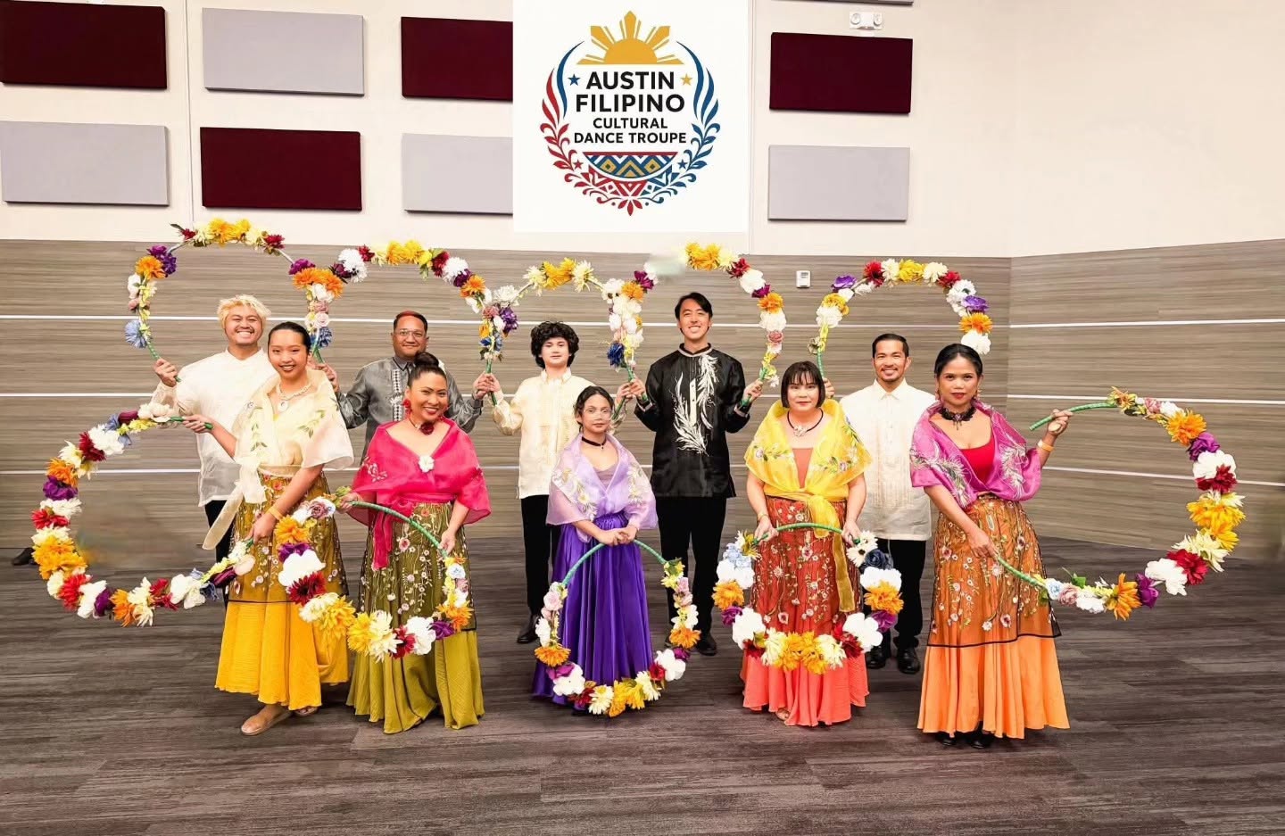 Group of nine people in colorful traditional costumes on stage with floral decorations and a sign reading 'Austin Filipino Cultural Dance Troupe'.