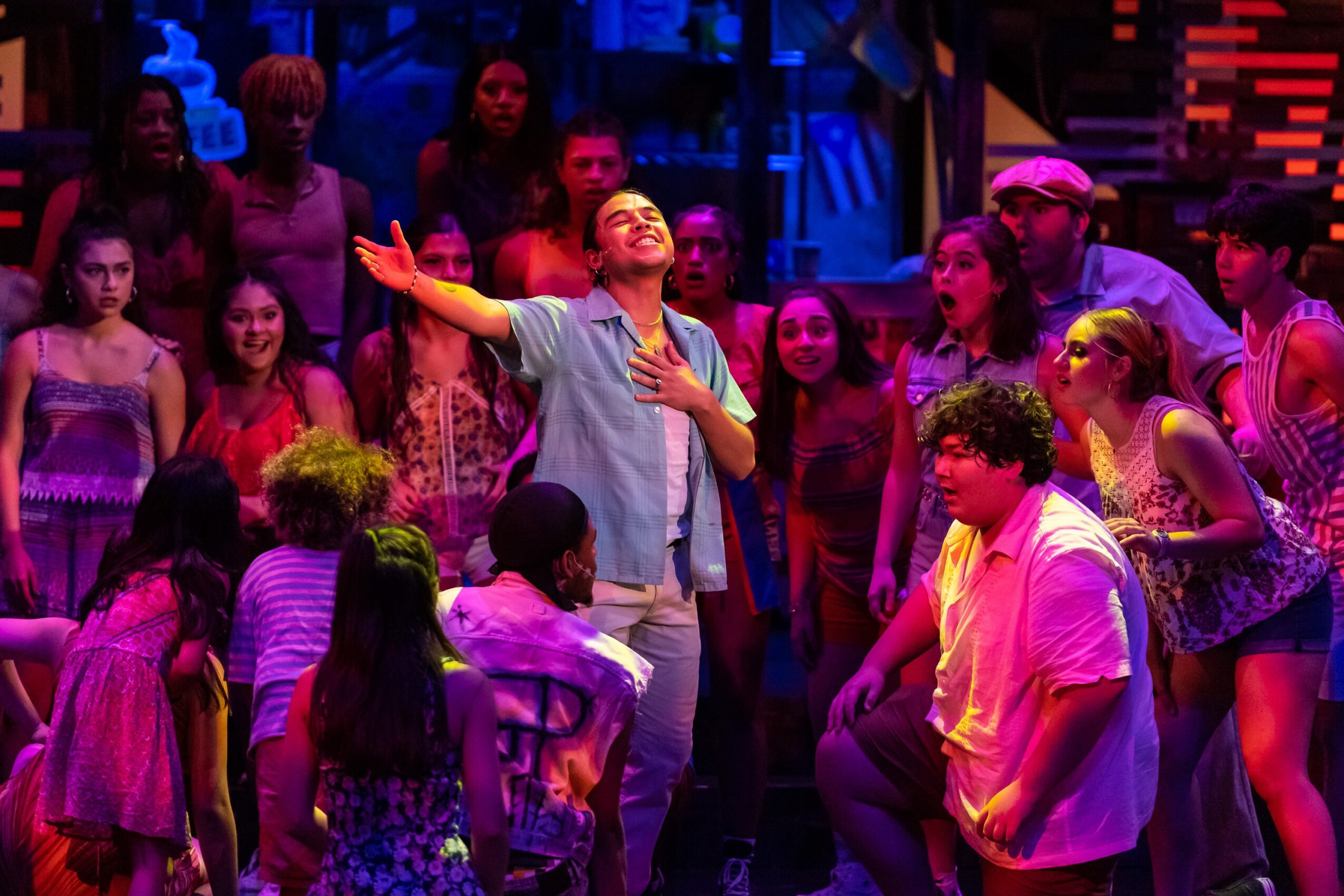 A performer sings center stage as a colorful group of people surrounds him under blue and pink lighting.