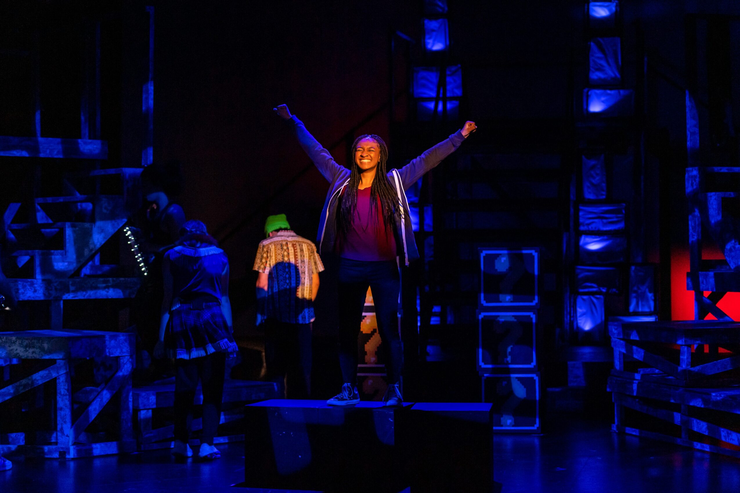 A person stands on a raised platform with arms lifted, surrounded by performers and stage equipment under blue lighting.