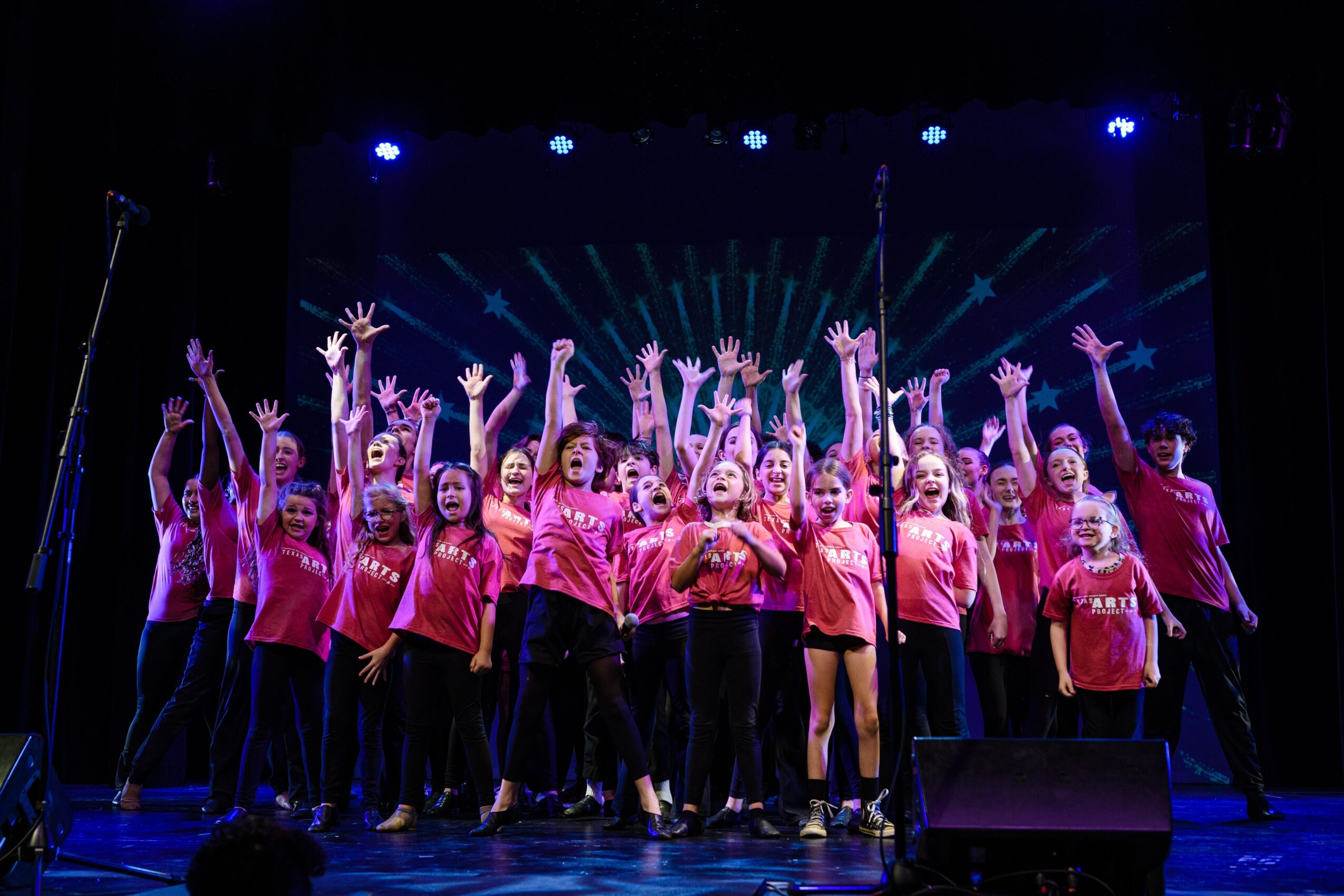 A group of children and adults in matching shirts stand onstage with their arms raised beneath colorful lights.