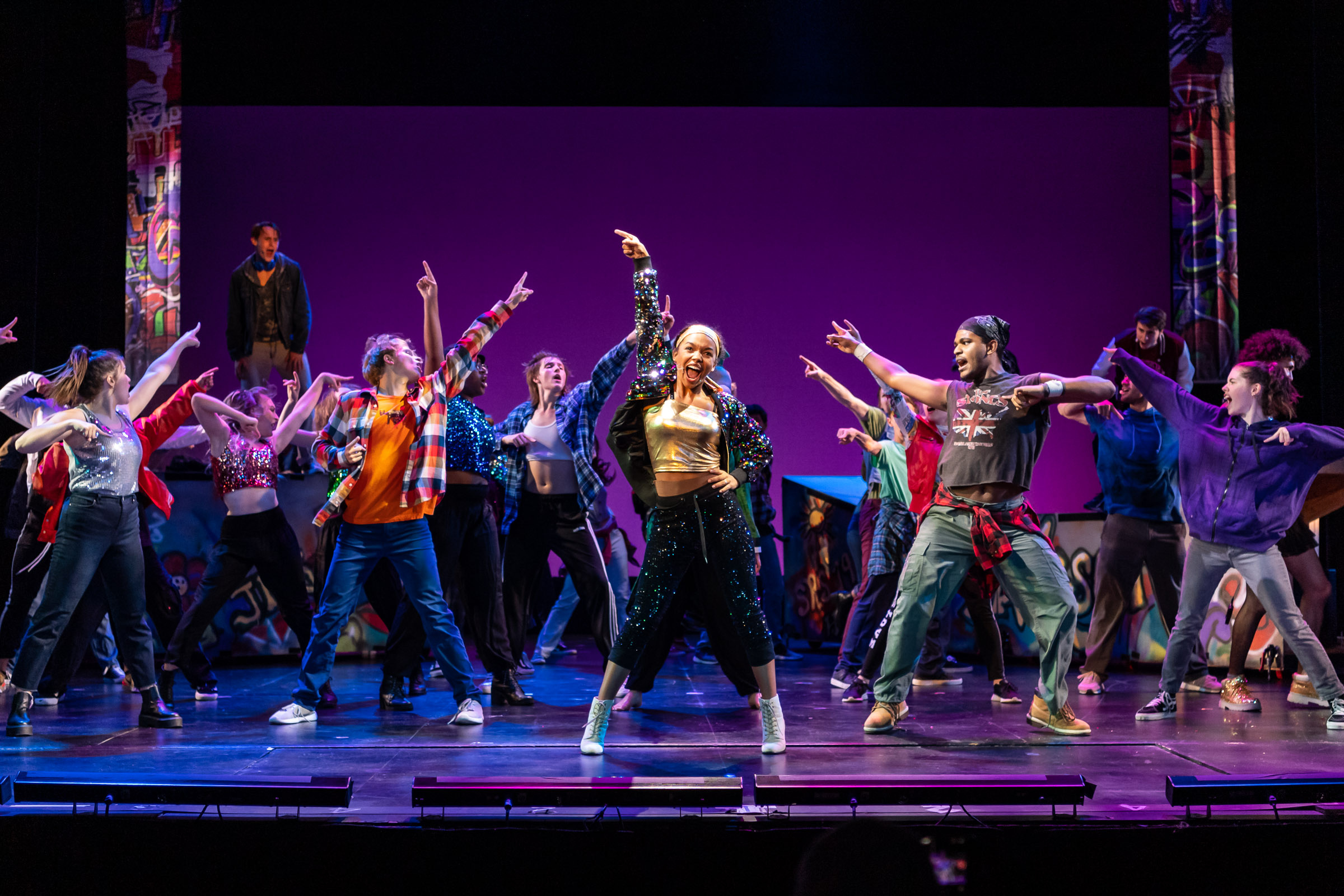 A group of performers dance across a stage under purple lighting, with a central performer holding a microphone.