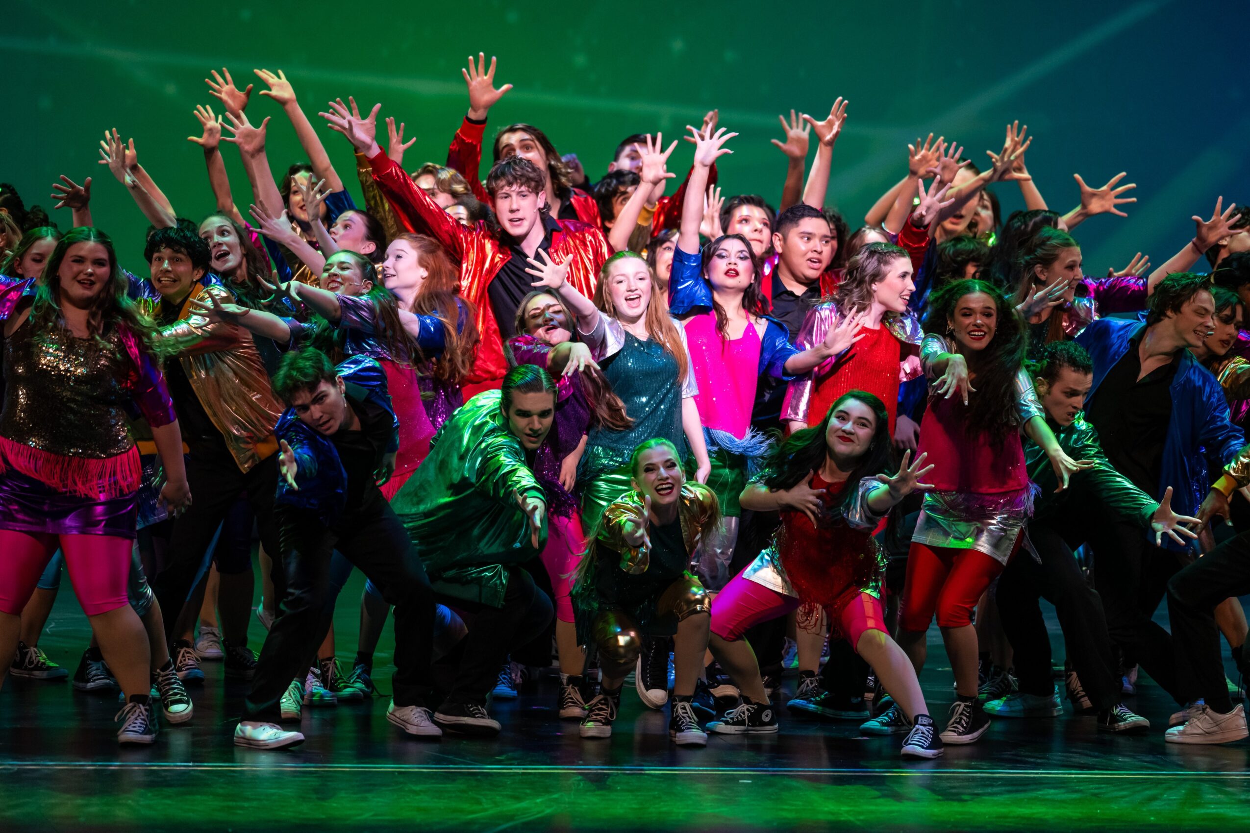 A large group of performers in colorful costumes pose with raised arms on a stage under green lighting.
