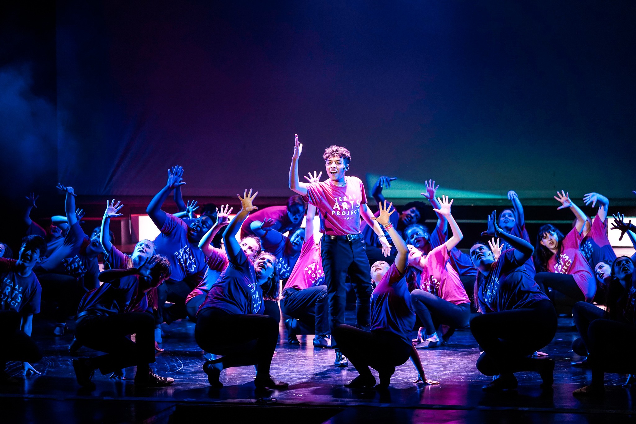 A performer stands center stage as a group of dancers crouch and reach around them under blue and pink lights.