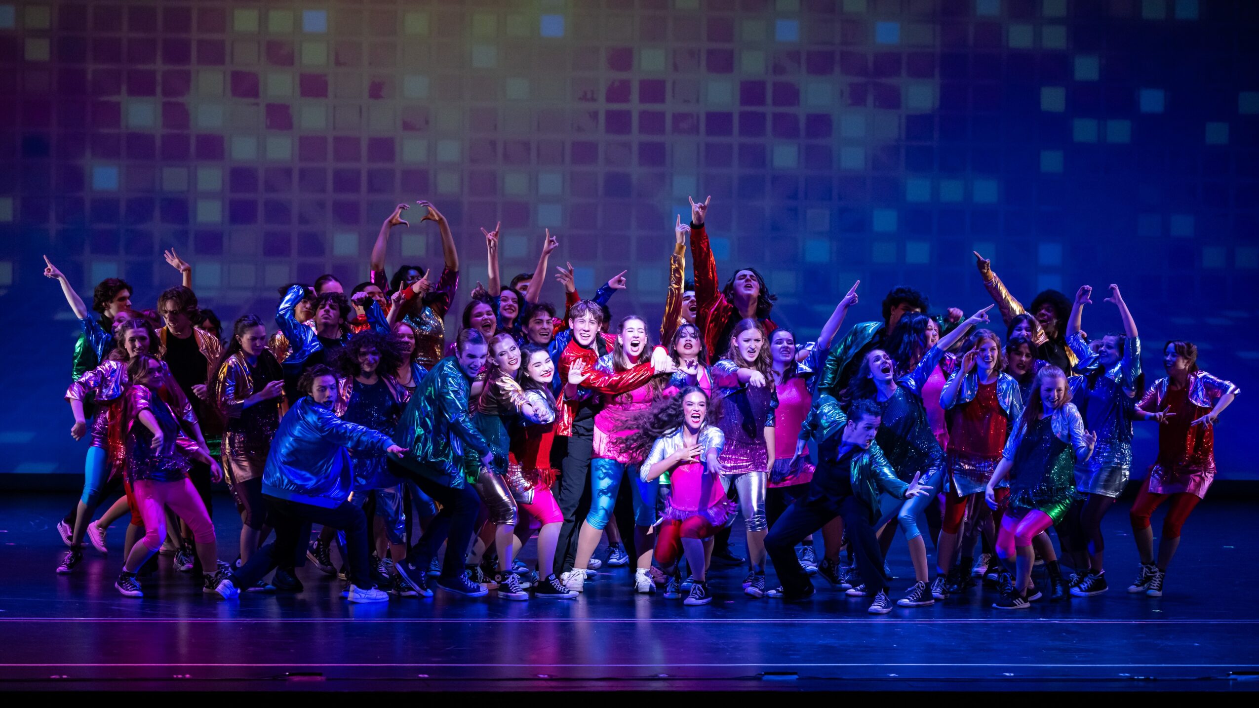 A large group of performers poses on a stage under colorful lights.