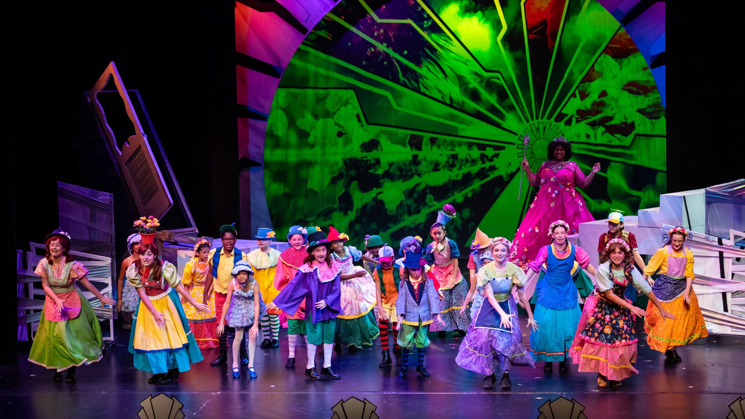 A colorful stage production with a large ensemble in bright costumes beneath a vivid green circular backdrop.