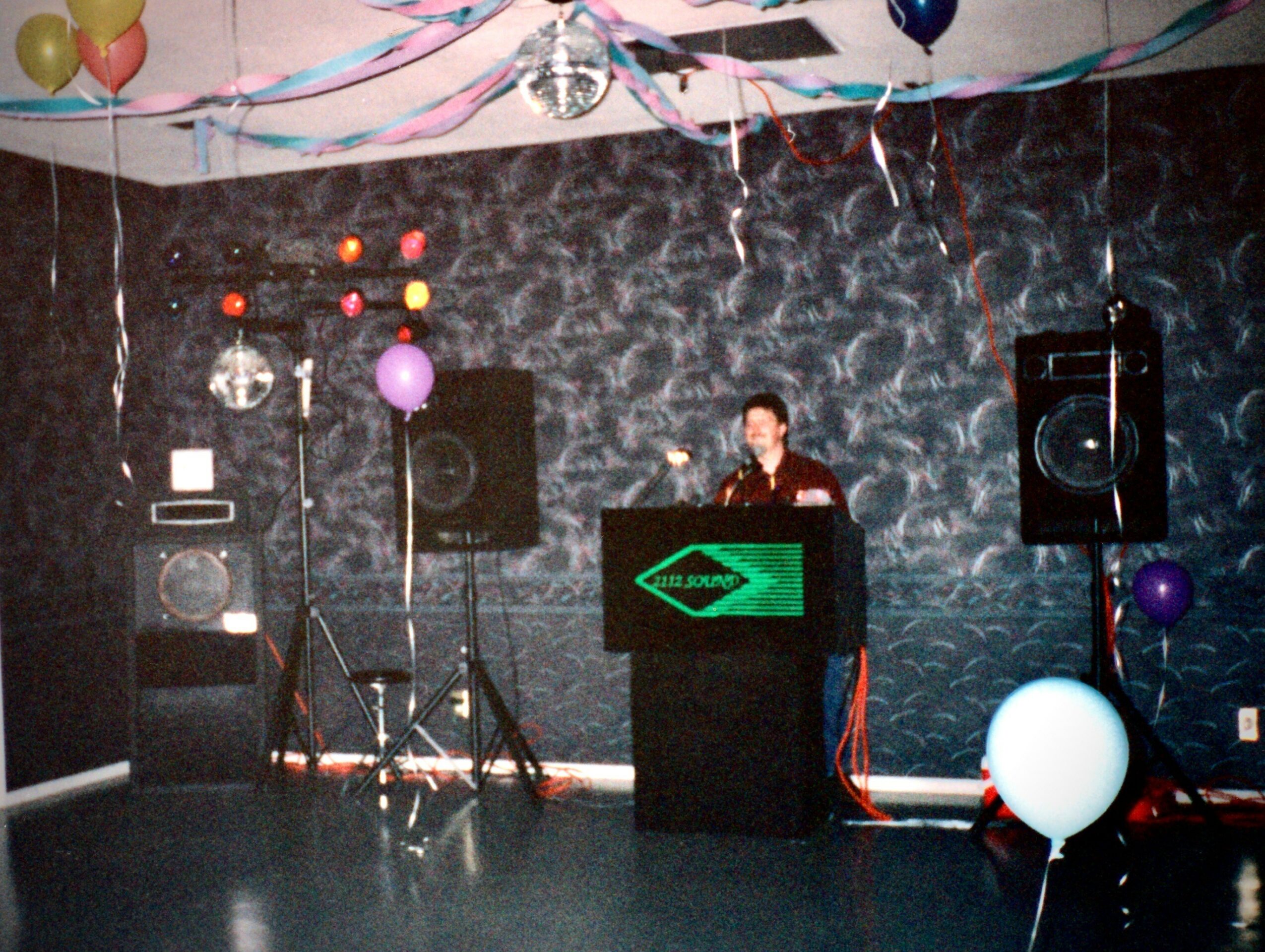 Person DJing at a decorated party with balloons and streamers, black textured wall background, speakers, and a green-lit DJ booth.