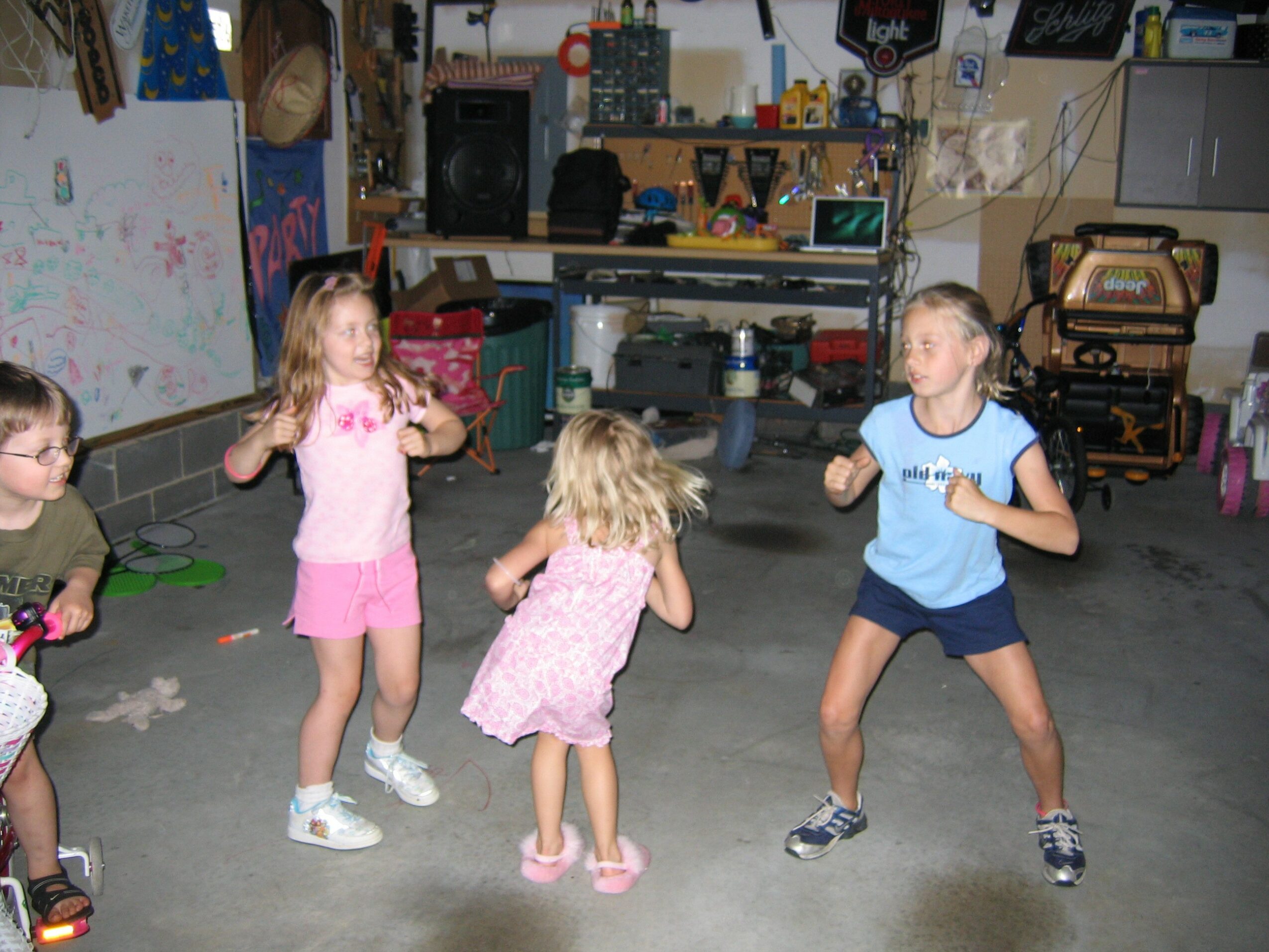 Four children dancing and playing in a garage with shelves, tools, and storage items visible in the background.