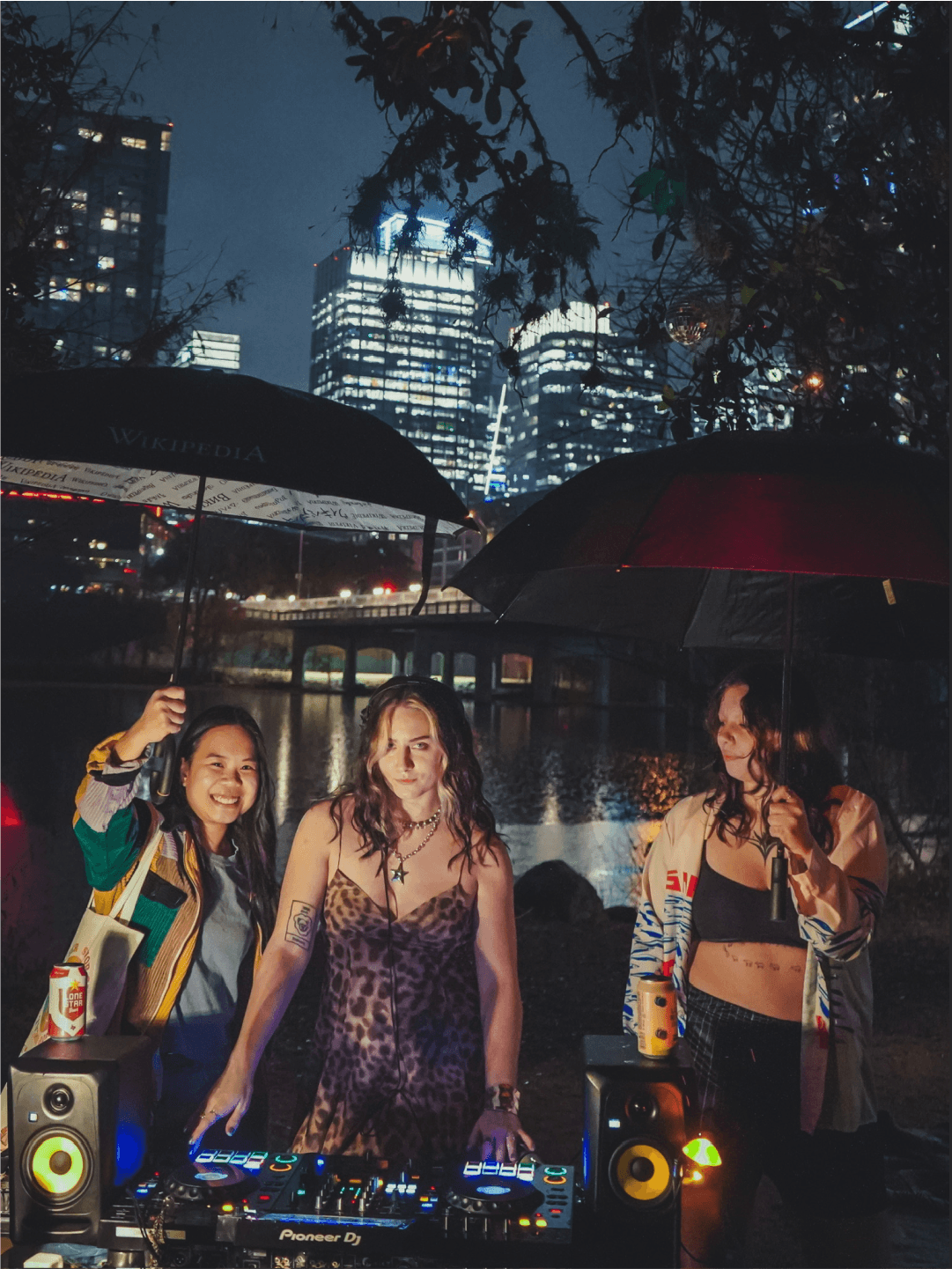 Three women stand outdoors at night with city skyline in background, holding umbrellas, near DJ equipment, smiling.