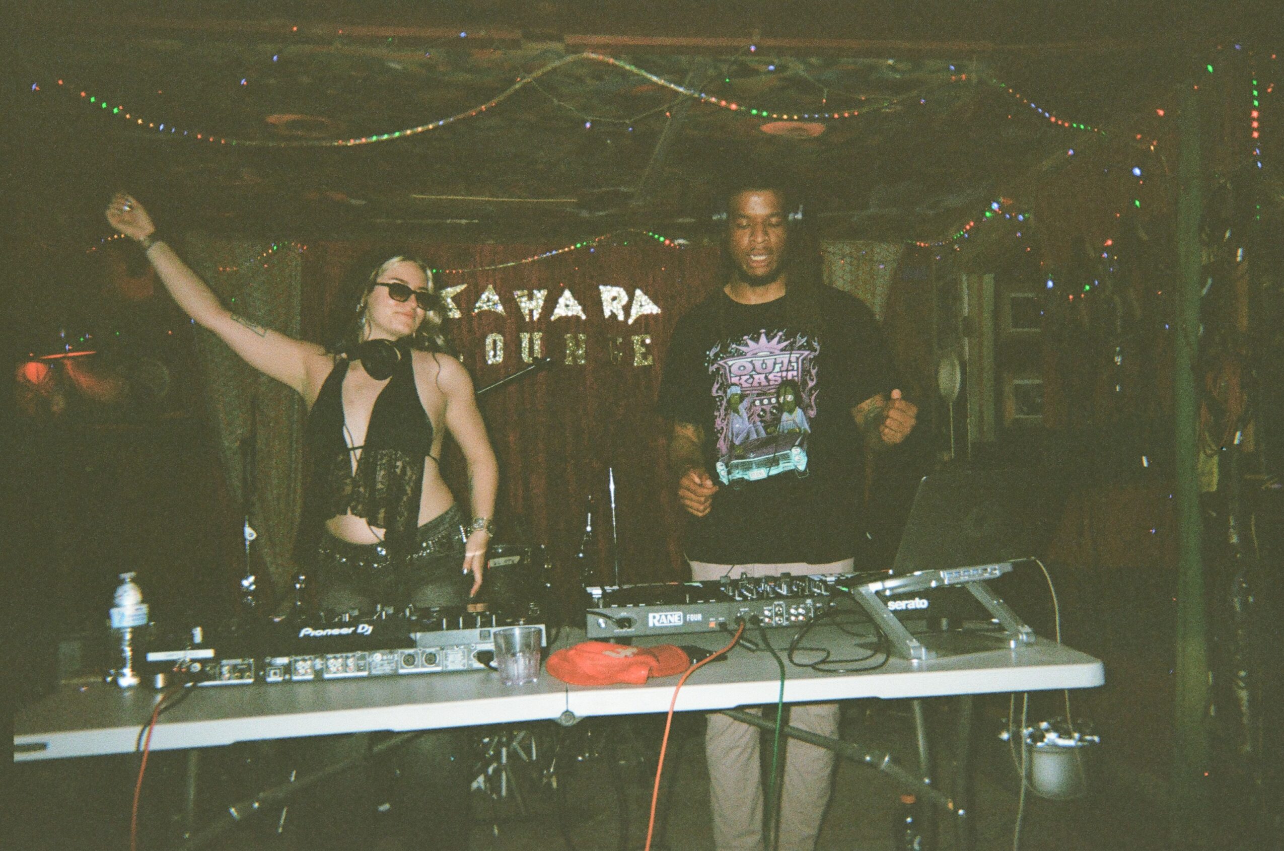 Two people stand behind DJ equipment in a dimly lit club with colorful lights and a sign in the background.