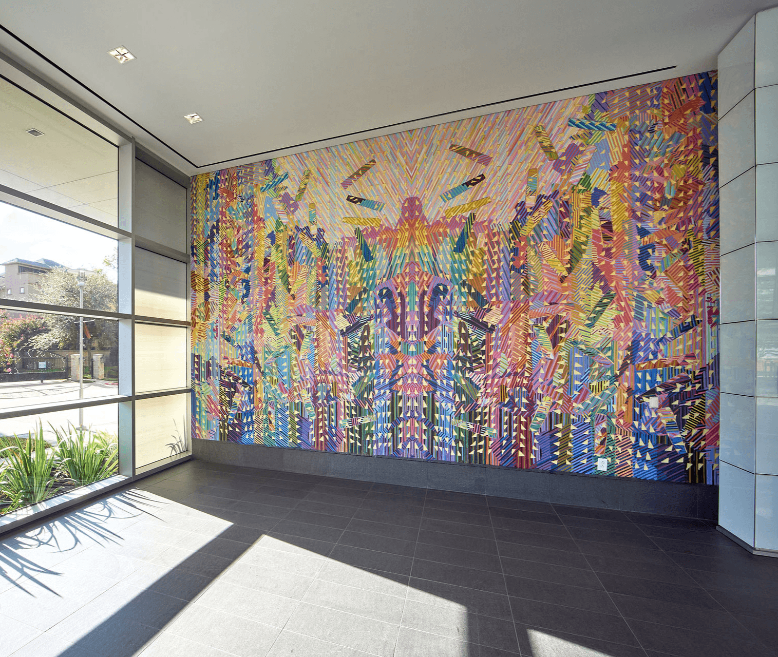 Colorful abstract mural on a wall inside a modern building with large windows and sunlight, tiled floor, and potted plants.