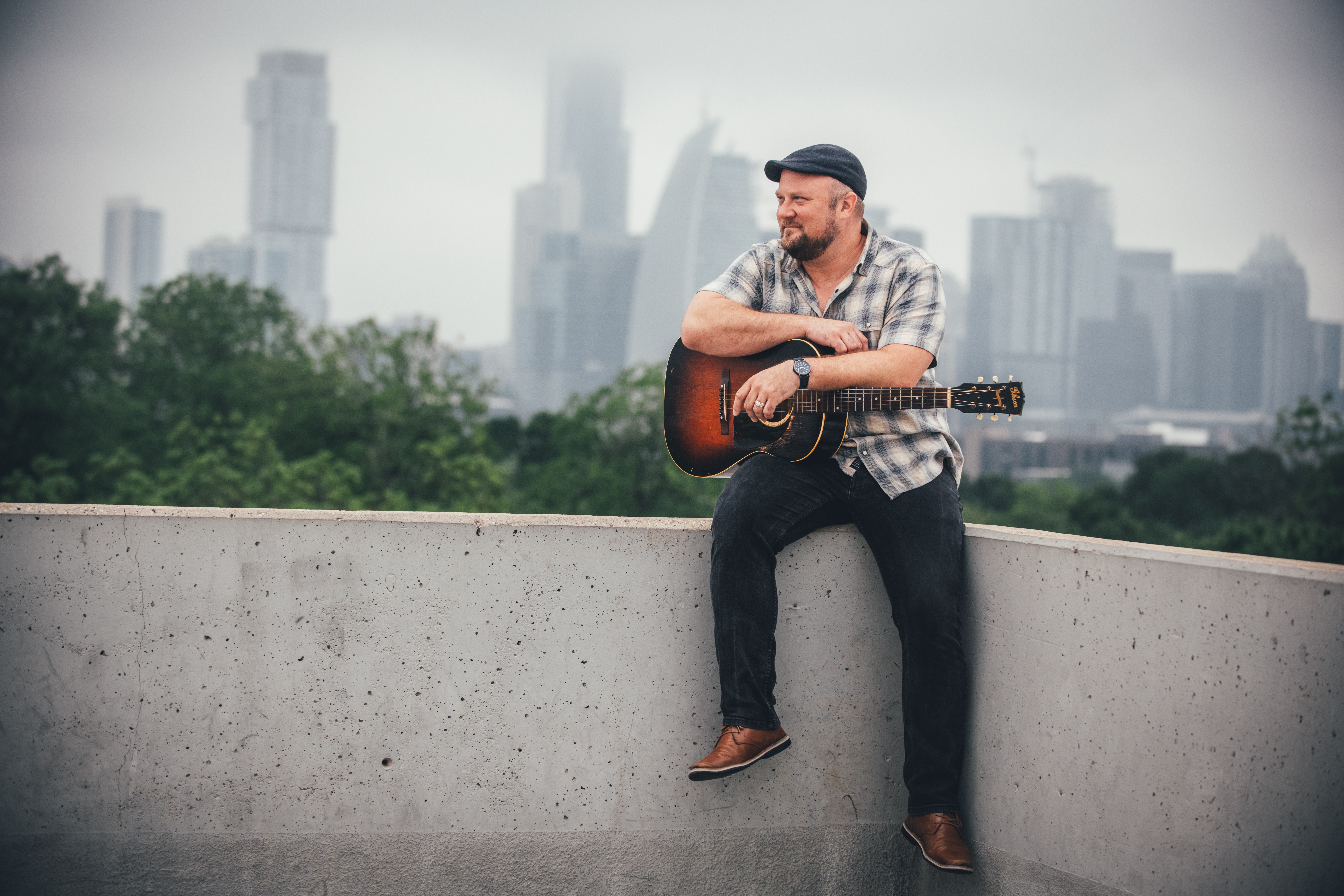 Man sitting on a concrete ledge with a guitar, city skyline in the background, overcast sky.