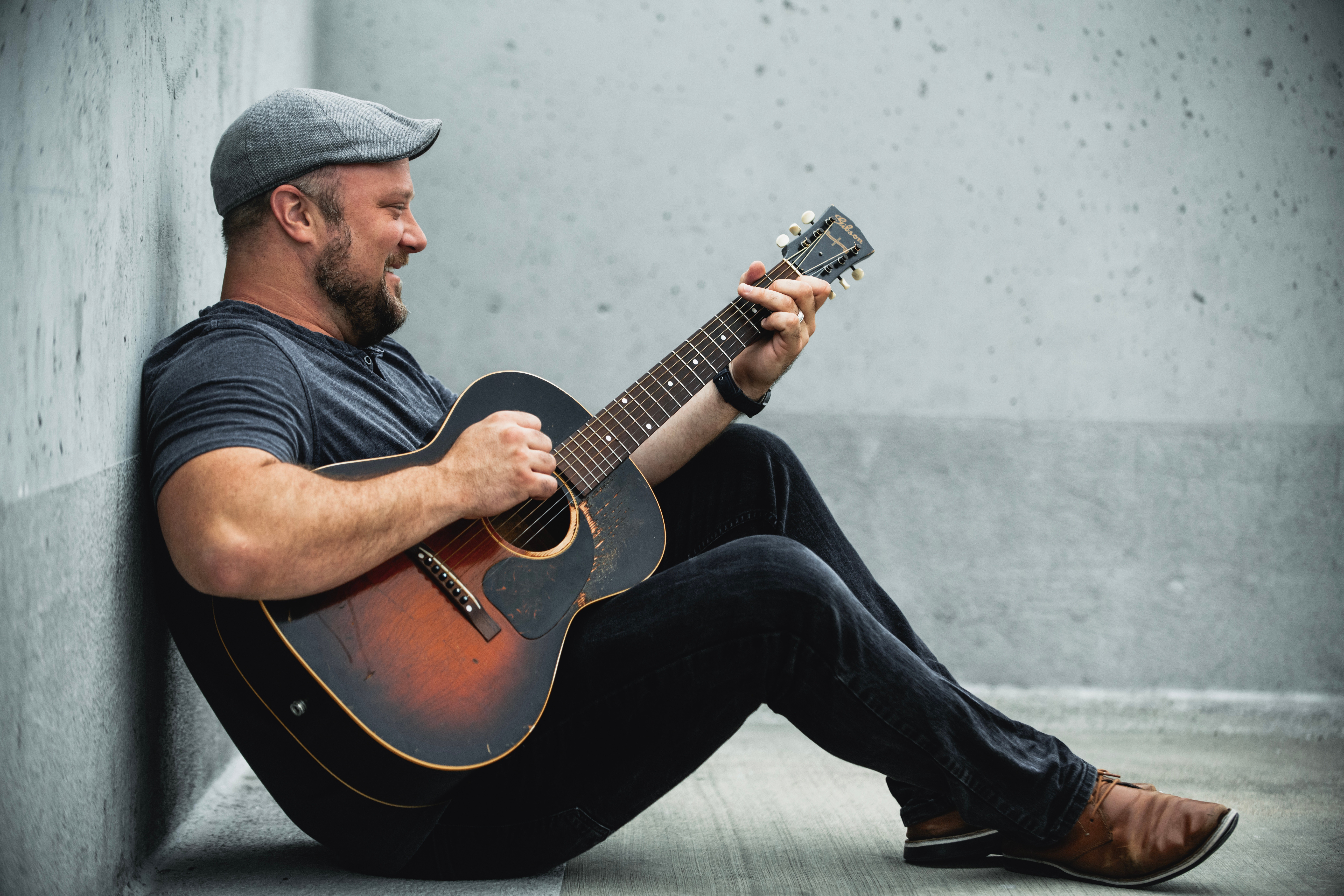 Man sitting on the floor playing an acoustic guitar, leaning against a wall, smiling, wearing a cap and casual clothes.