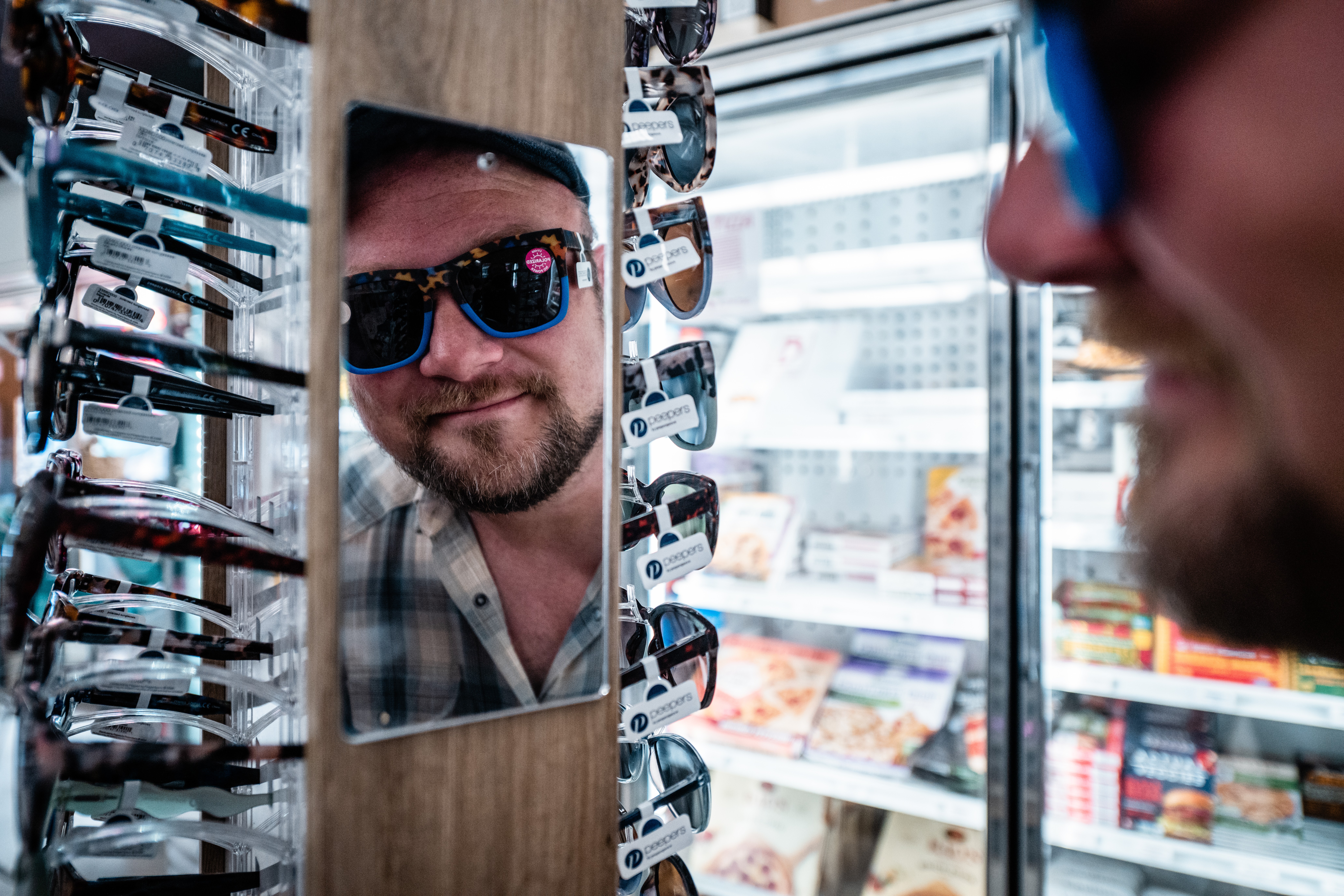 Man wearing sunglasses looking at his reflection in a mirror surrounded by sunglasses display, store interior visible in background.