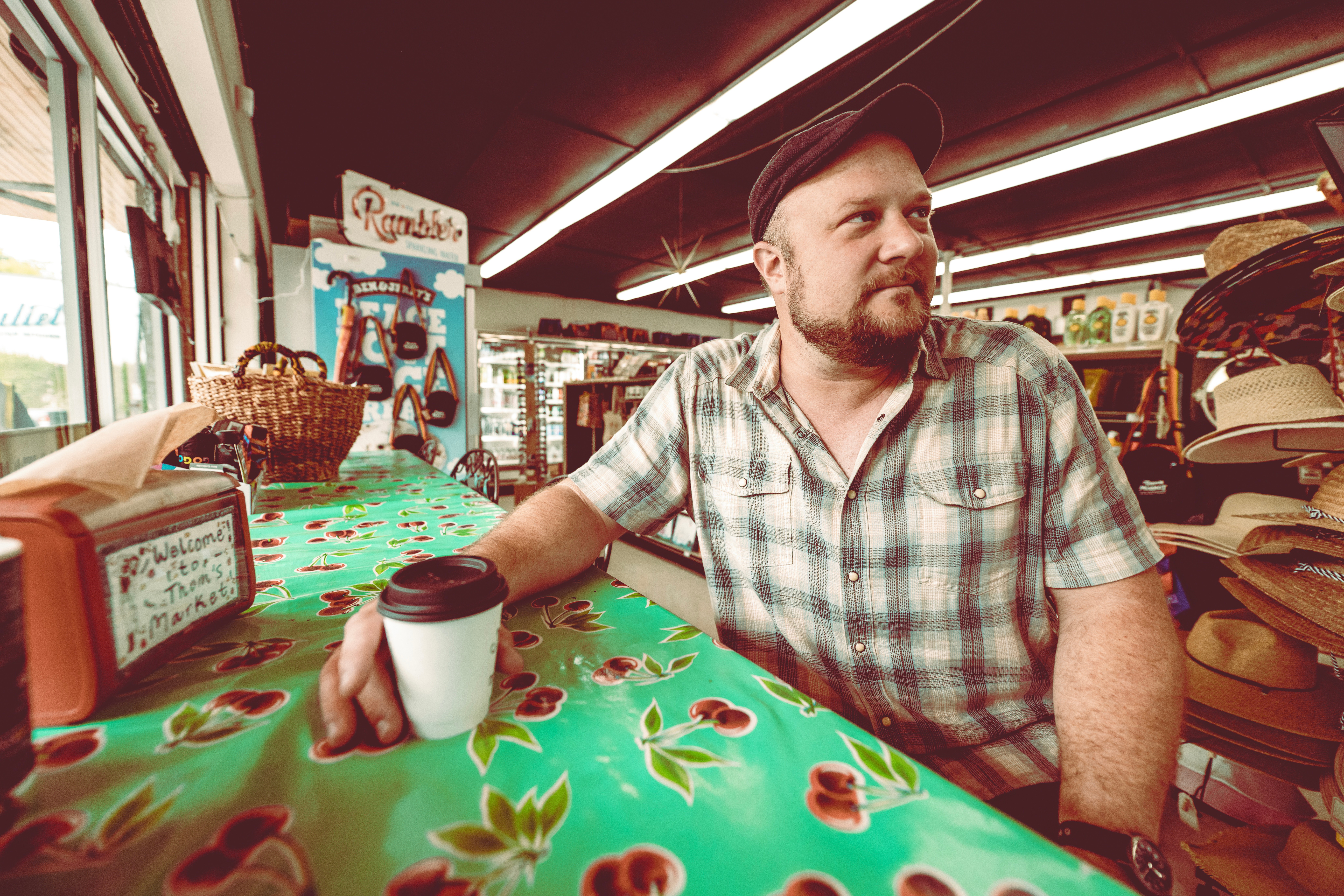Man with beard in plaid shirt sitting at a store counter with hats on shelves behind him, holding a coffee cup, looking to the side.