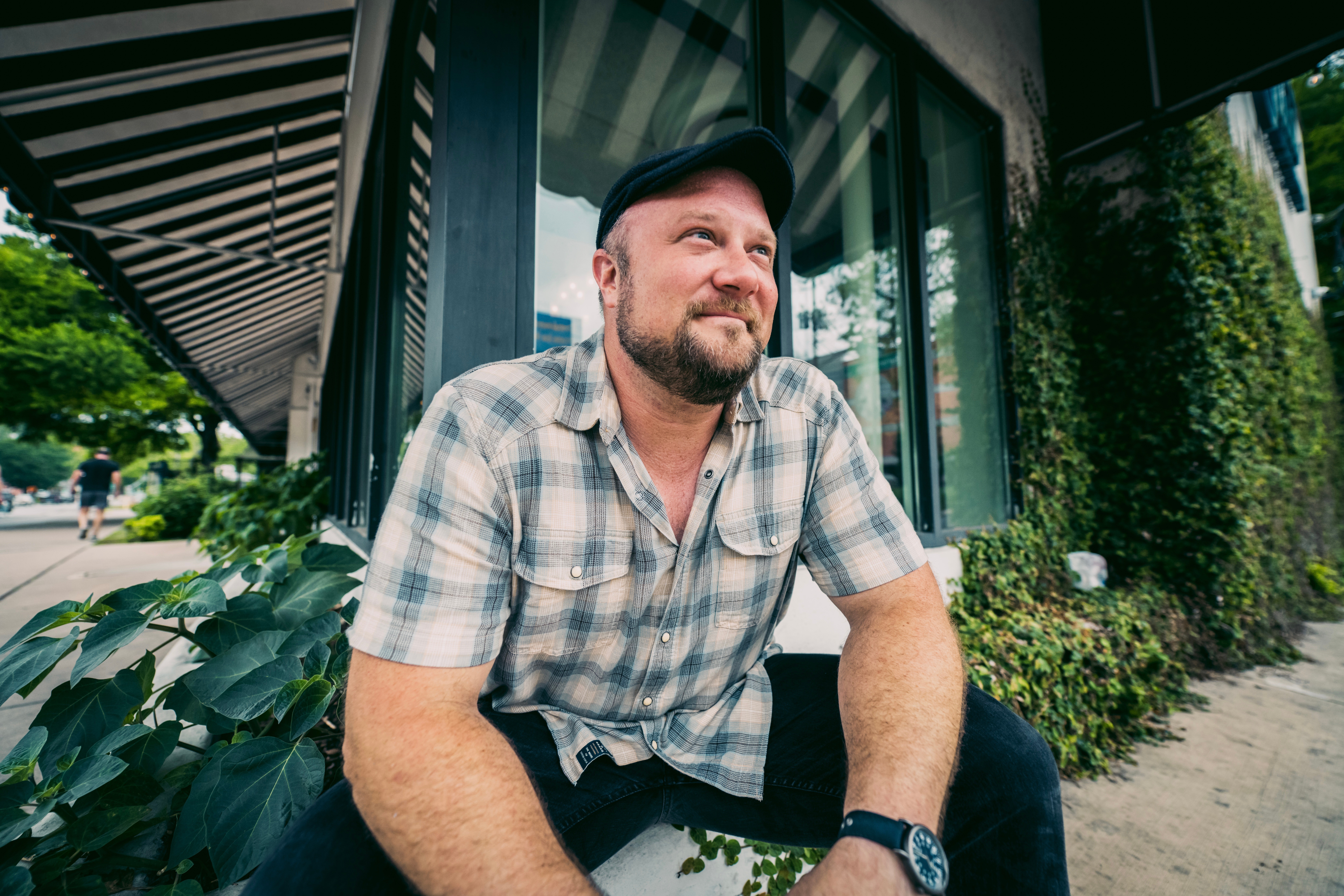 Man with a beard wearing a plaid shirt and cap sitting outdoors near a building with greenery.