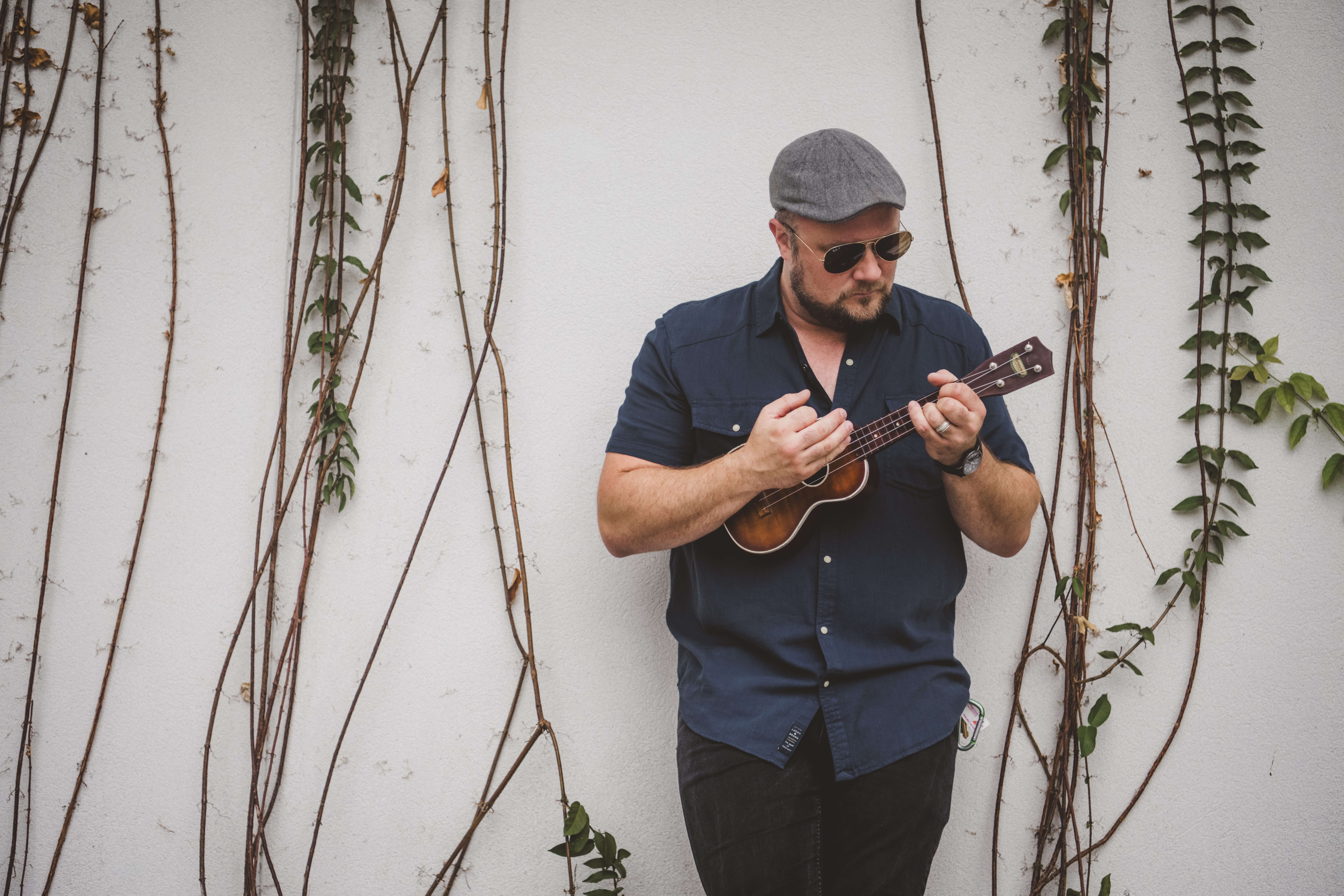 Man wearing sunglasses, a gray cap, and dark clothing playing a ukulele against a white wall with vines.