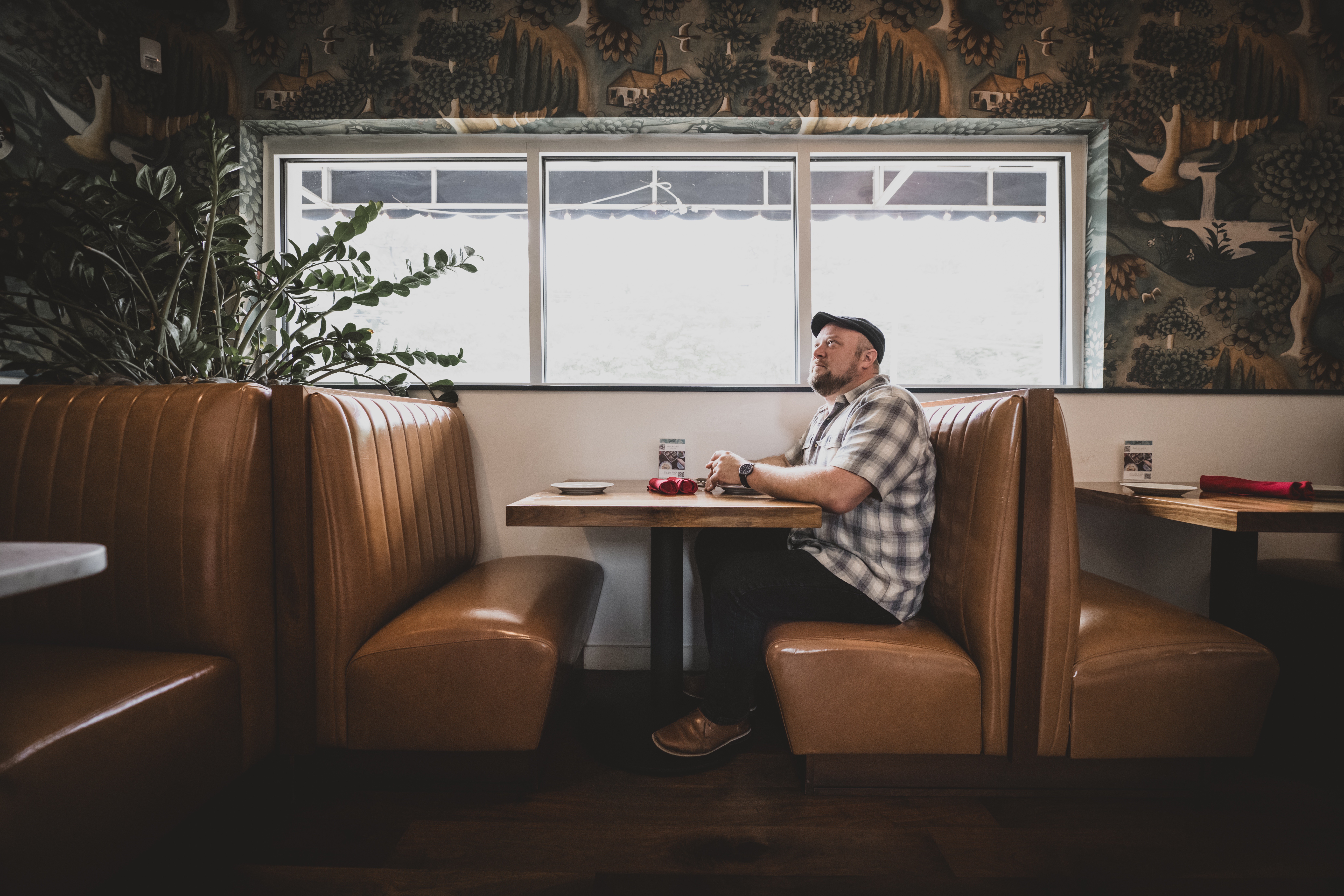 Person sitting alone at a booth in a restaurant, looking out the window, with a plant nearby and a mural on the wall.