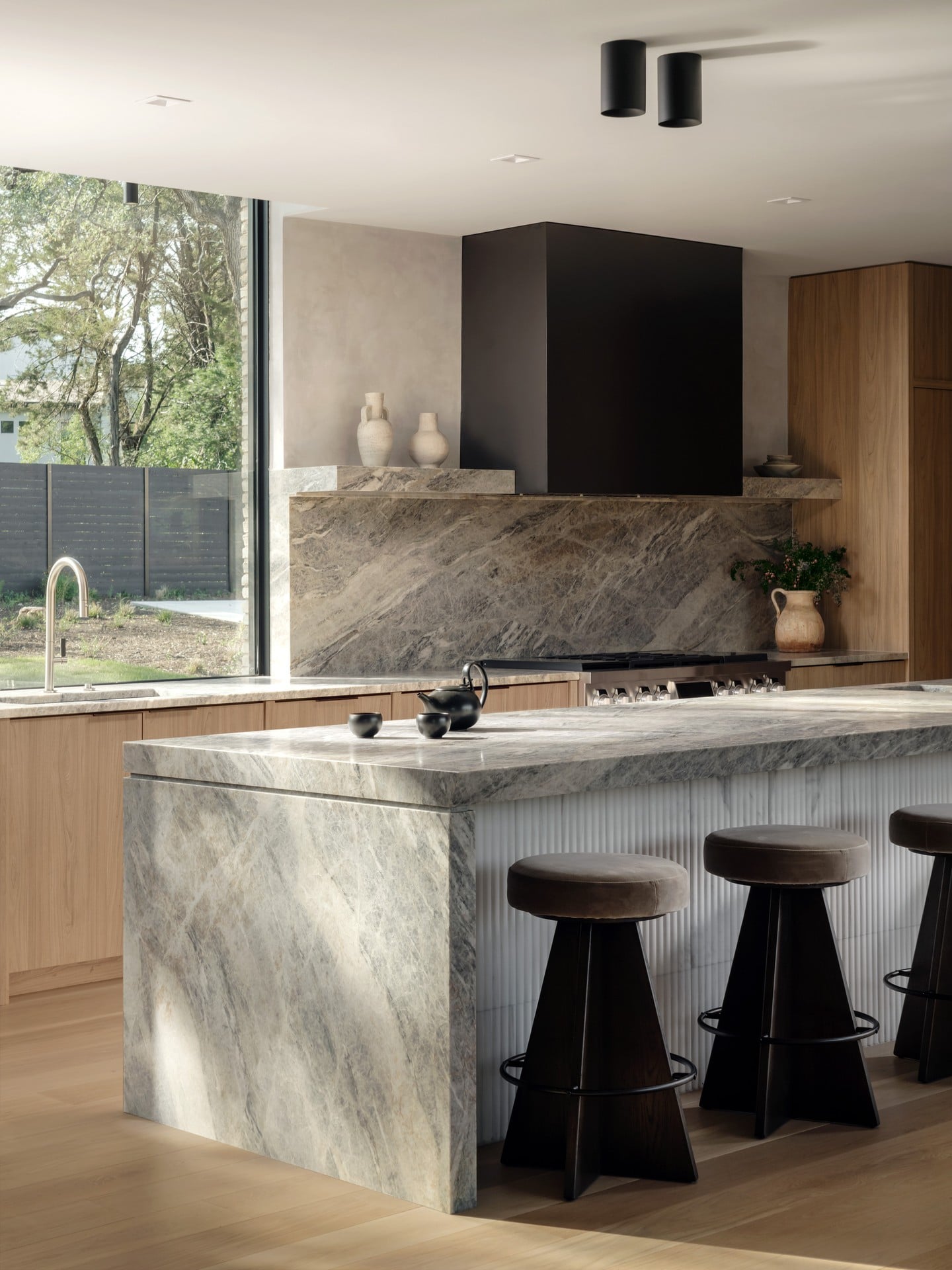 Modern kitchen with marble island, black stools, and large window showing outdoor trees.