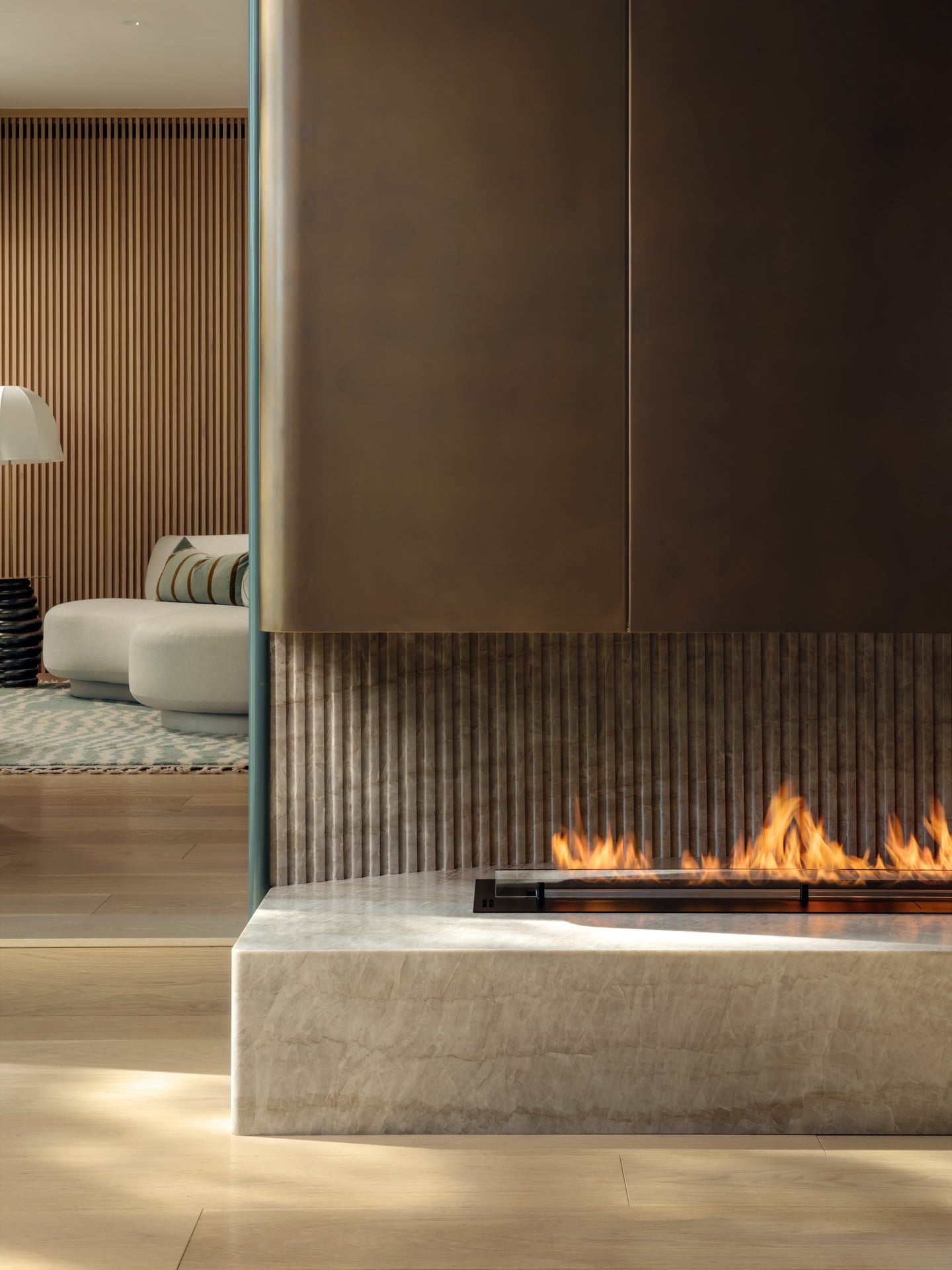Modern fireplace with flames, part of a room with dark wall and textured paneling, adjacent to a living area with sofa and wooden accents.