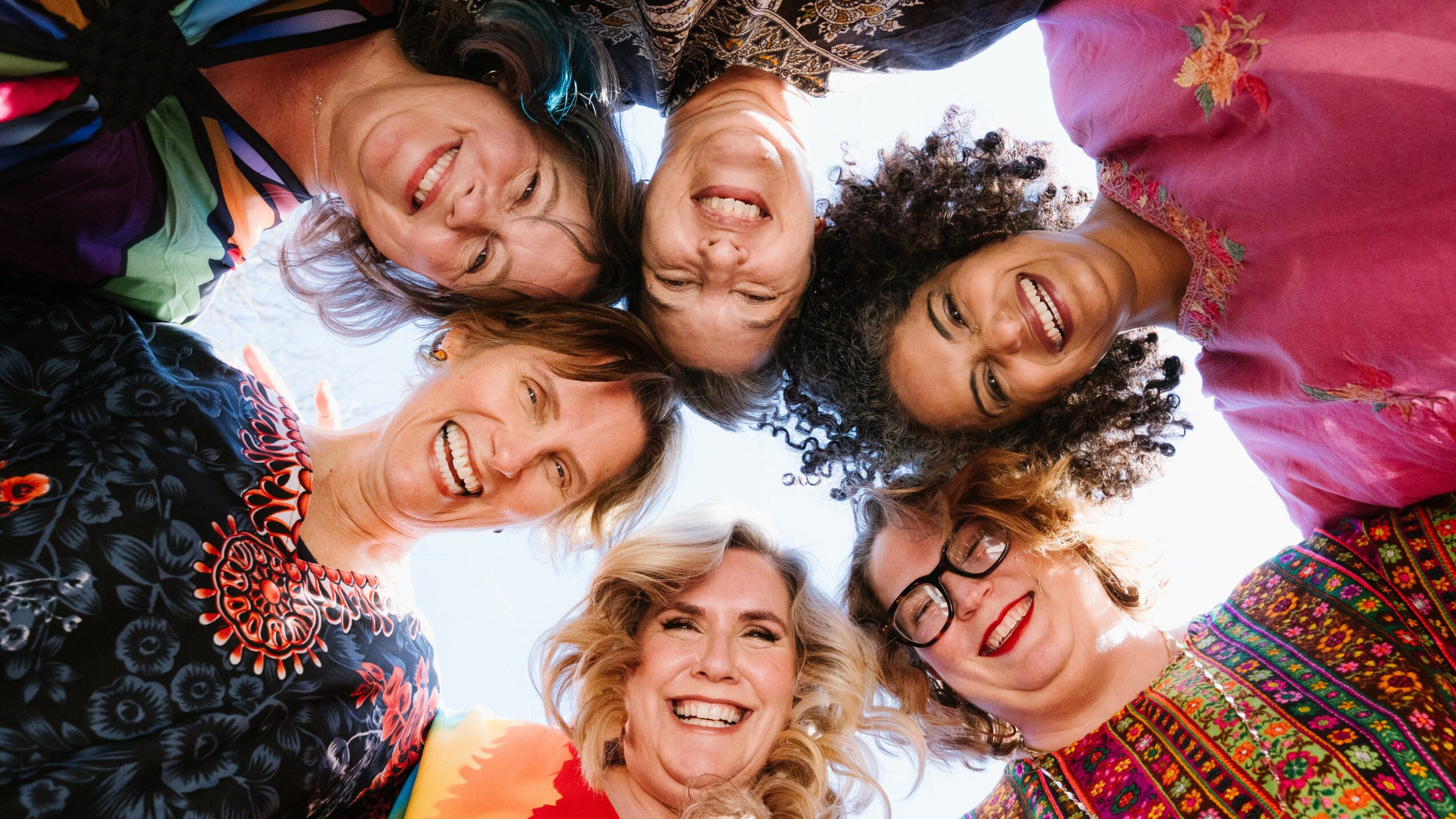 Six women in colorful clothing smiling and looking down at the camera in a circle outdoors.