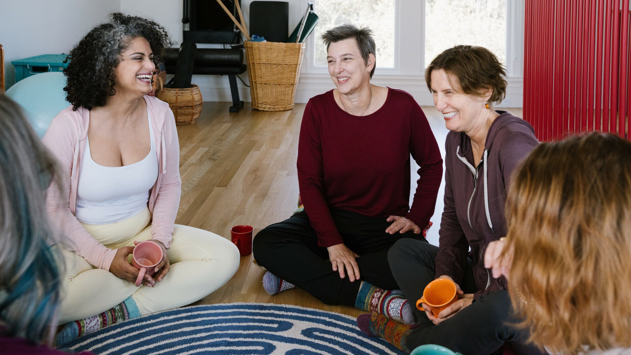 Four women sitting on the floor in a circle, smiling and talking in a cozy room.