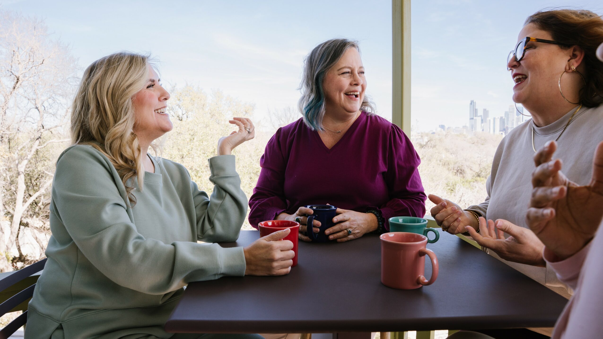 Four women sitting at a table, smiling and talking, with a window and outdoor scenery in the background.