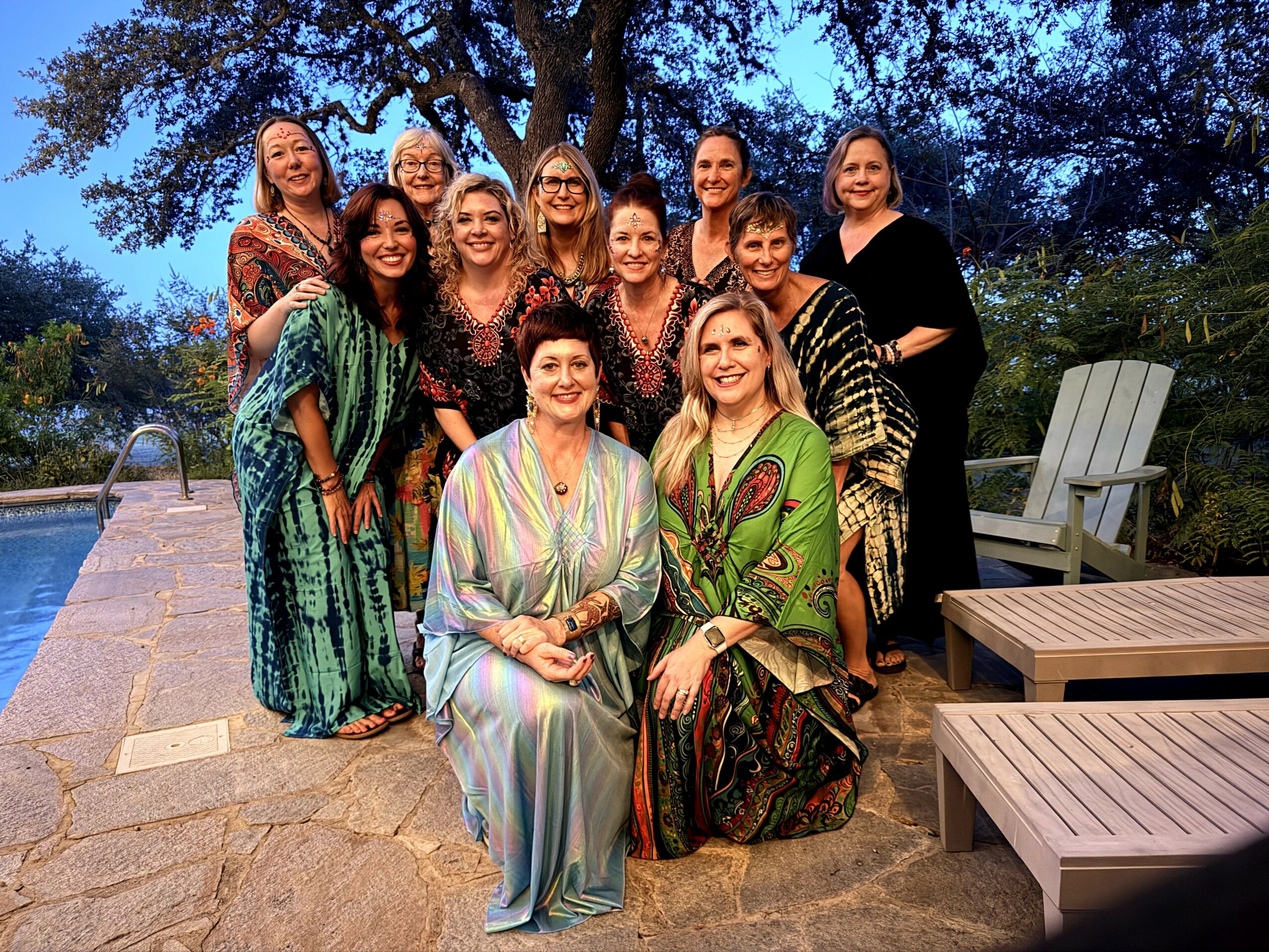 Group of ten women outdoors near a pool, some sitting and some standing, with trees and evening sky in background.