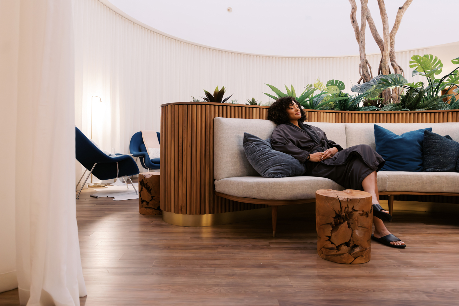 Person reclining on a sofa in a lounge with plants, wooden furniture, and chairs.