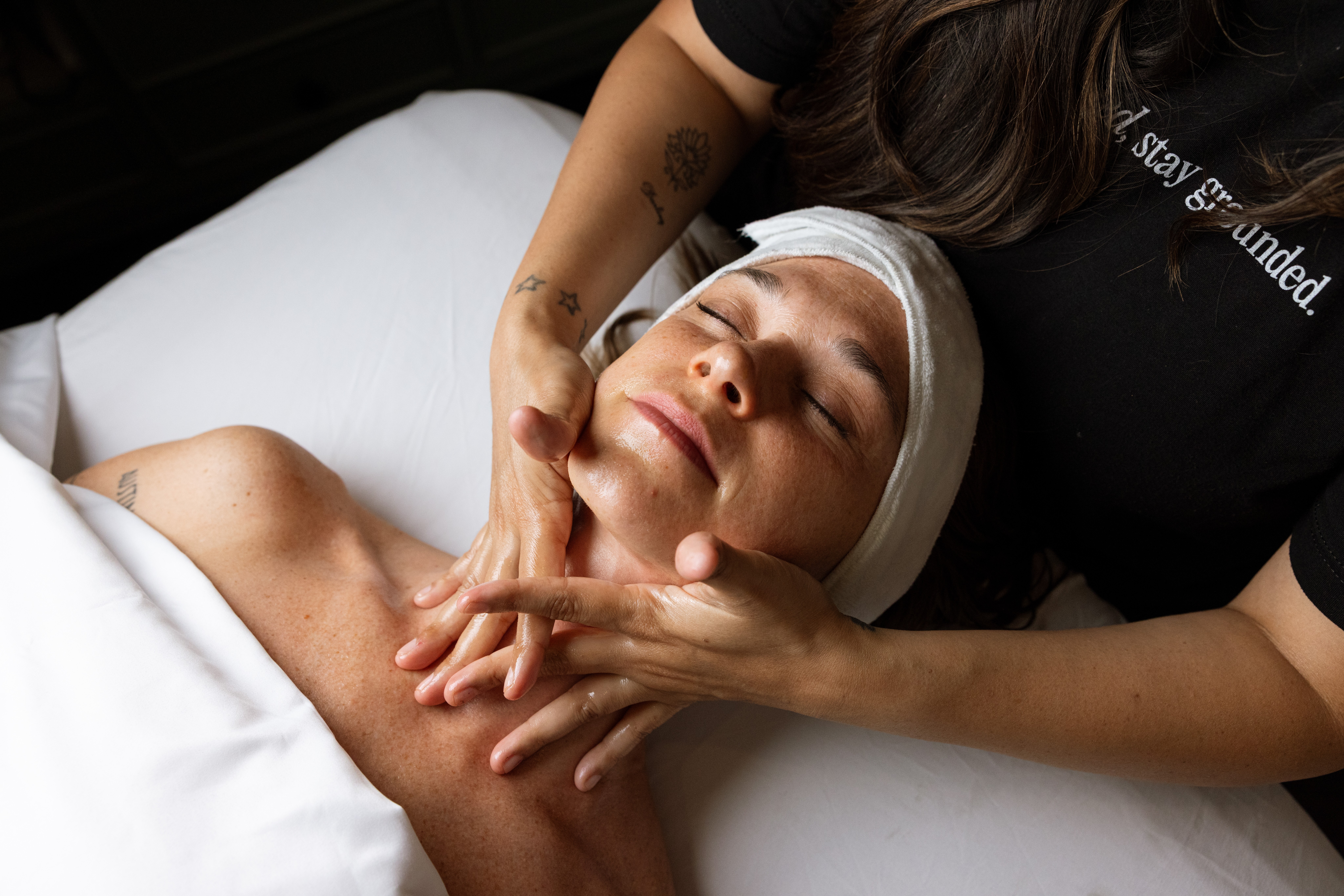 A person receives a facial massage while lying beneath a white sheet, with a practitioner holding their face.