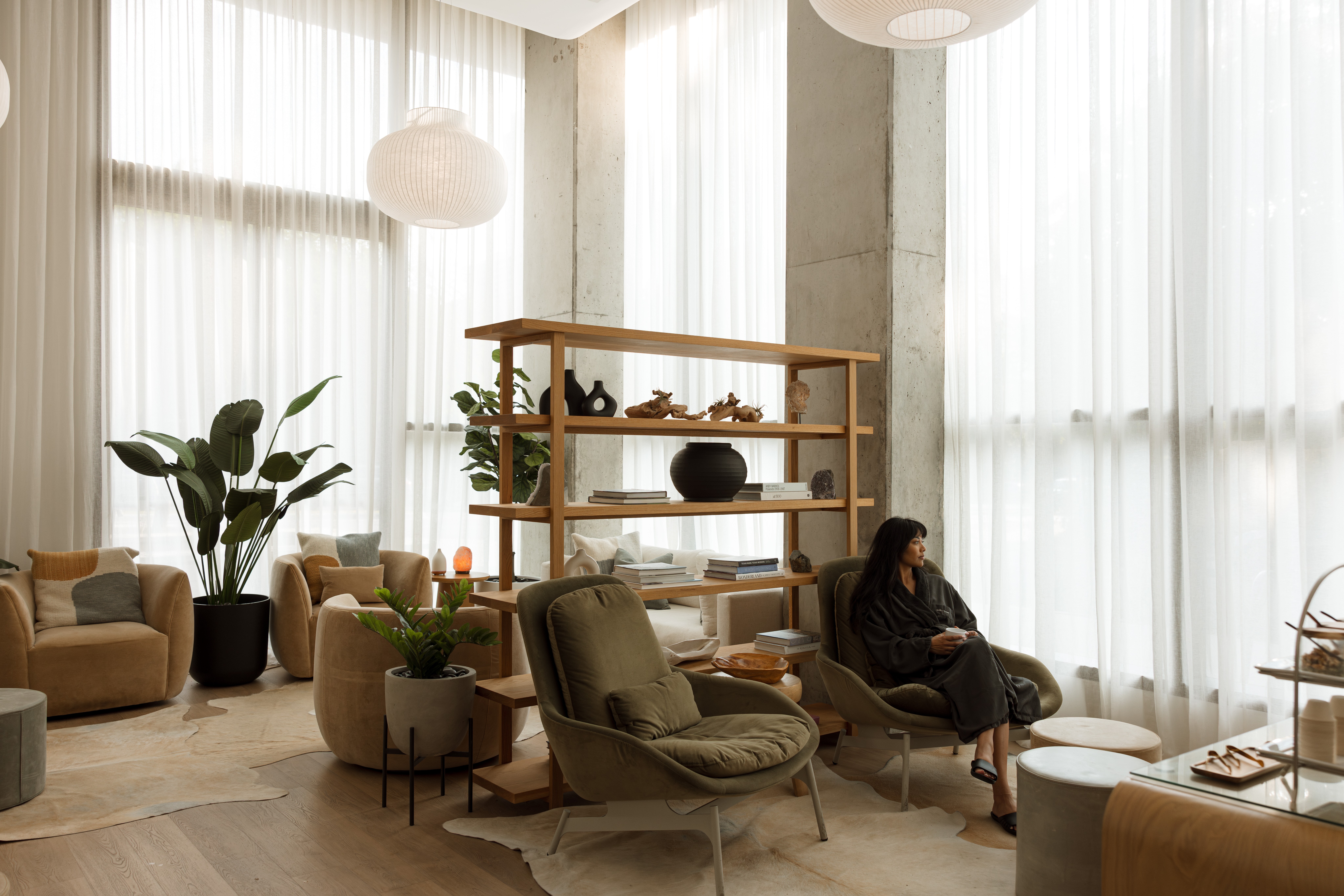 A bright living room with floor-to-ceiling curtains, lounge chairs, plants, and a wooden shelving unit.