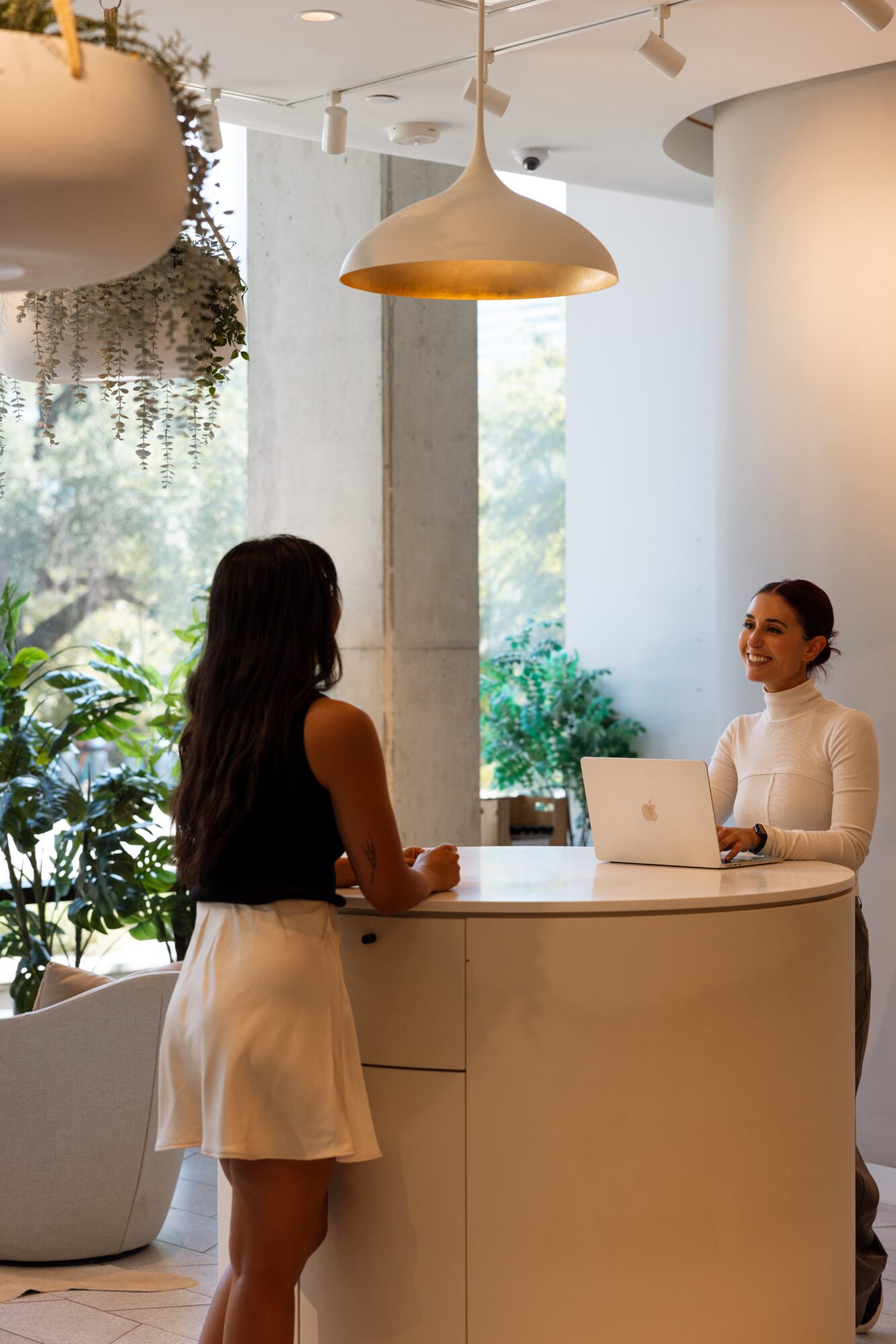 Two people stand at a curved reception desk beneath pendant lights, with plants and windows behind them.