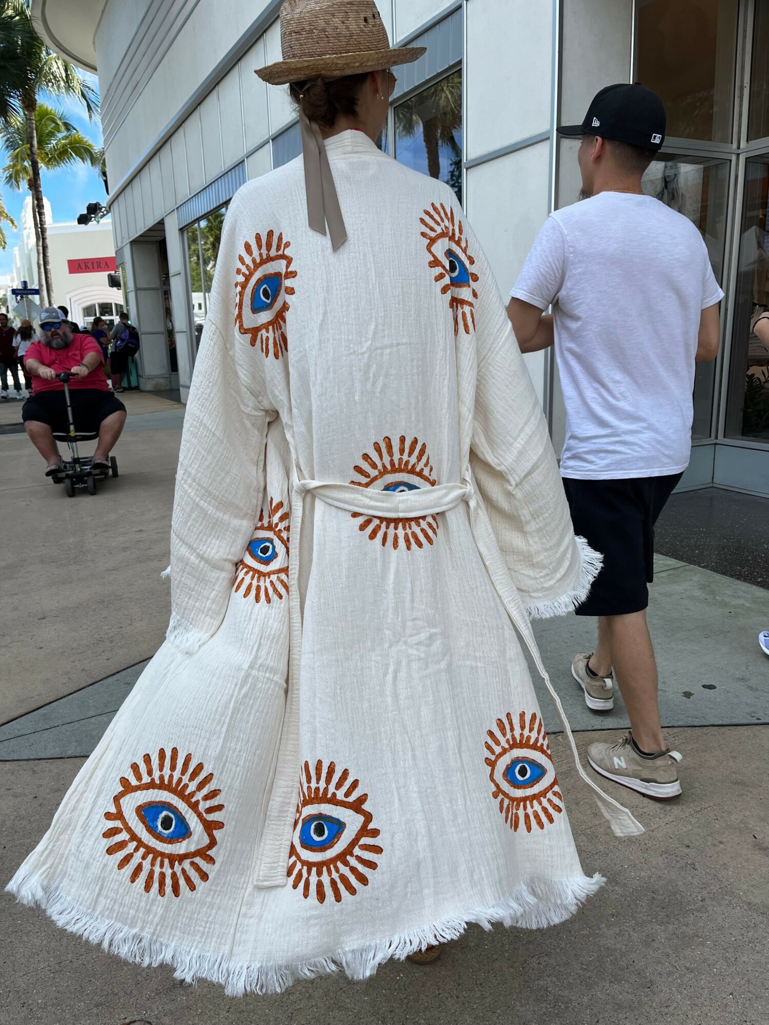 Person wearing a long white dress with eye designs, standing outdoors on a sidewalk.